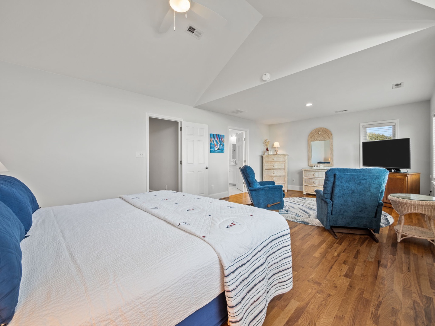 Relax in your spacious bedroom featuring vaulted ceilings, comfortable blue seating, and warm hardwood floors for the perfect coastal retreat.