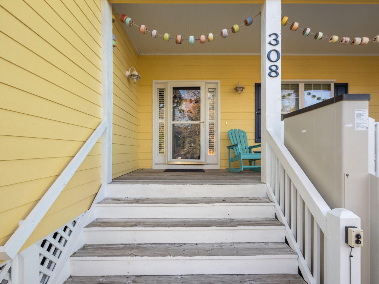 Charming yellow cottage entrance welcomes you with cheerful bunting and a cozy turquoise chair on the front porch.