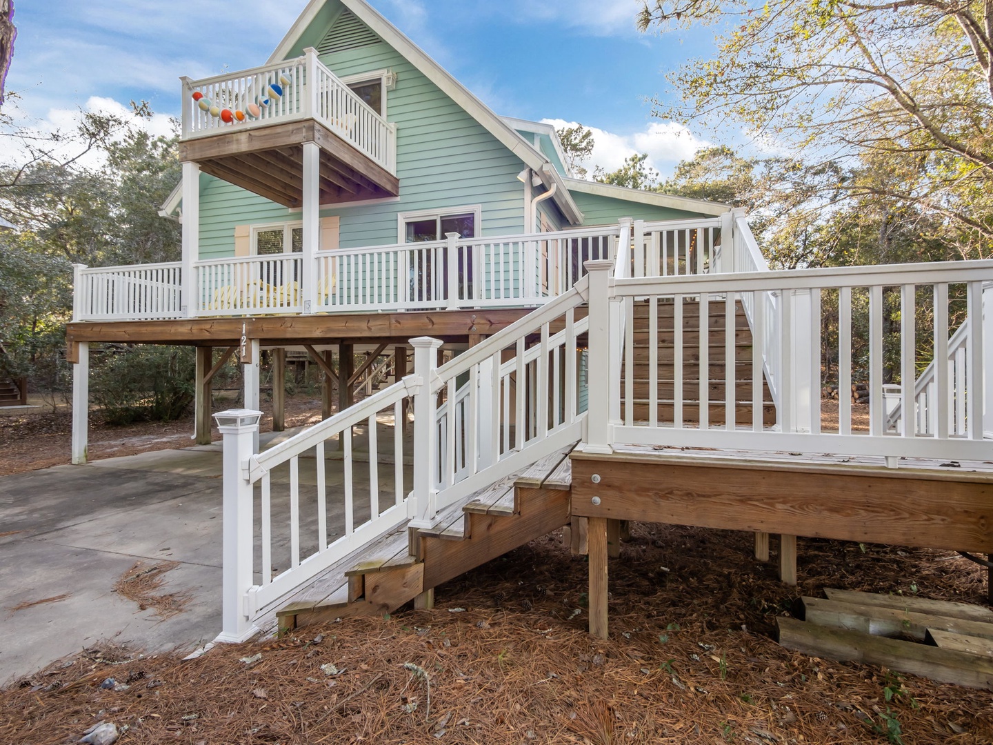 Charming raised beach house with multiple decks and staircases, nestled among mature trees in a peaceful wooded setting.