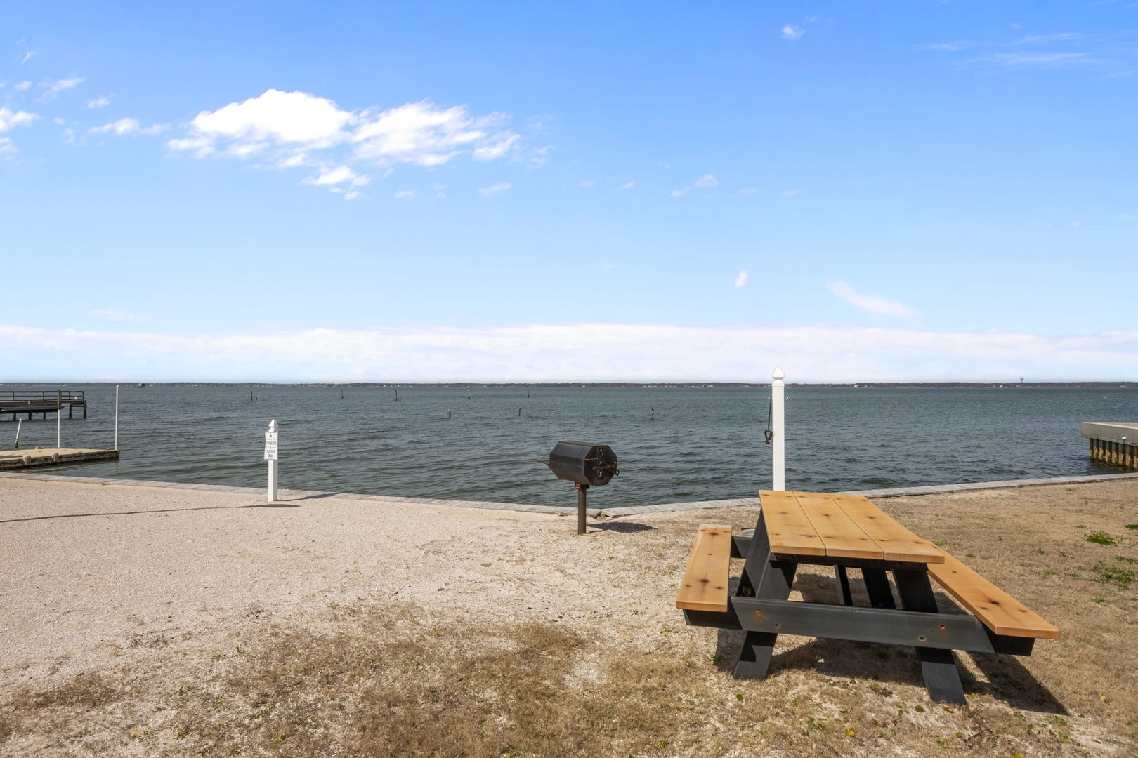 Waterfront picnic area with table and grill overlooking calm waters under blue skies.