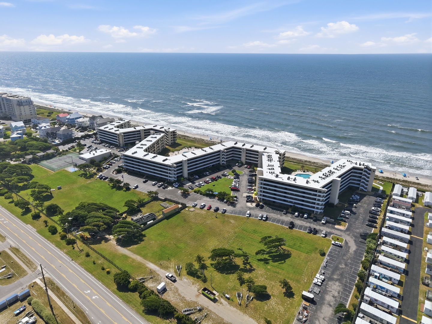 Aerial view of beachfront property with ocean access, green lawns, and modern multi-story buildings situated directly along the coast with rolling waves and sandy shoreline.