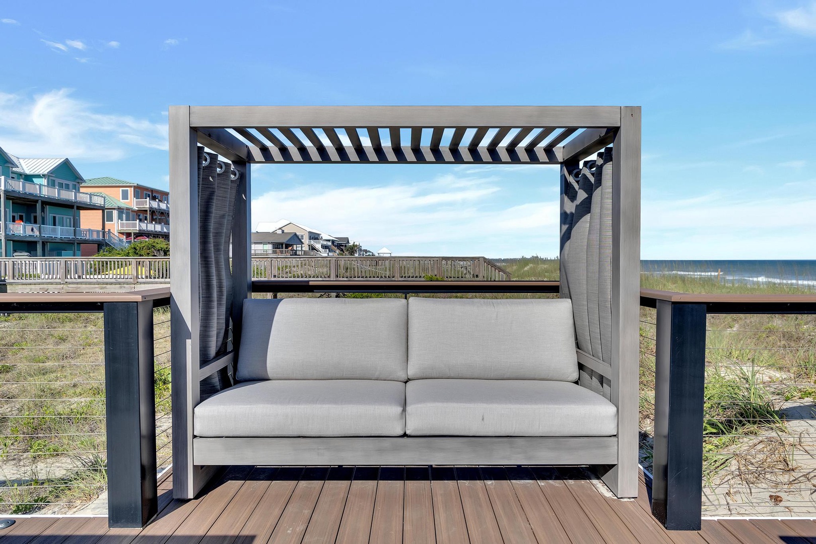 Oceanfront daybed with pergola shade on a private deck overlooking the beach and rolling waves, with direct beach access steps away.