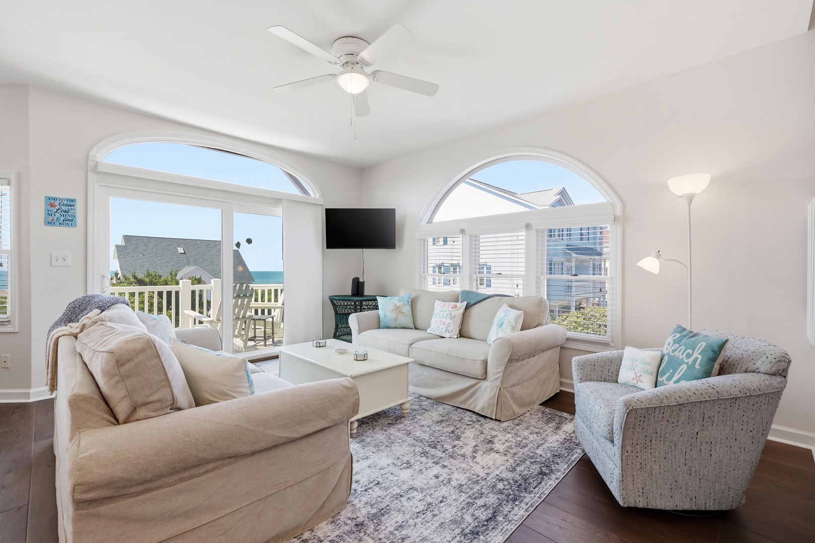 Unwind in your coastal sanctuary, where soft neutral tones meet ocean breezes. Picture yourself on plush seating, ocean views framed by elegant arched windows, ceiling fan spinning gently overhead.