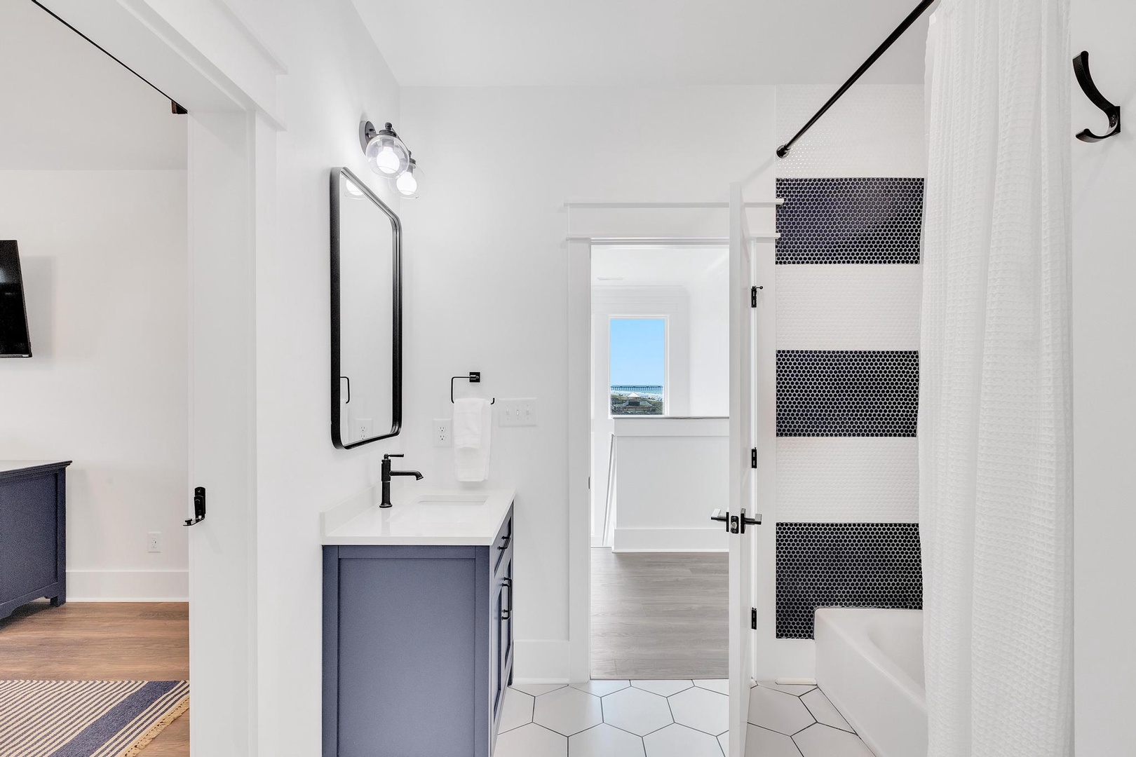 Your private bathroom features modern fixtures, sleek hexagon tile flooring, and a shower with striking black penny tile accents. Natural light flows through the adjacent room, adding warmth to the space.
