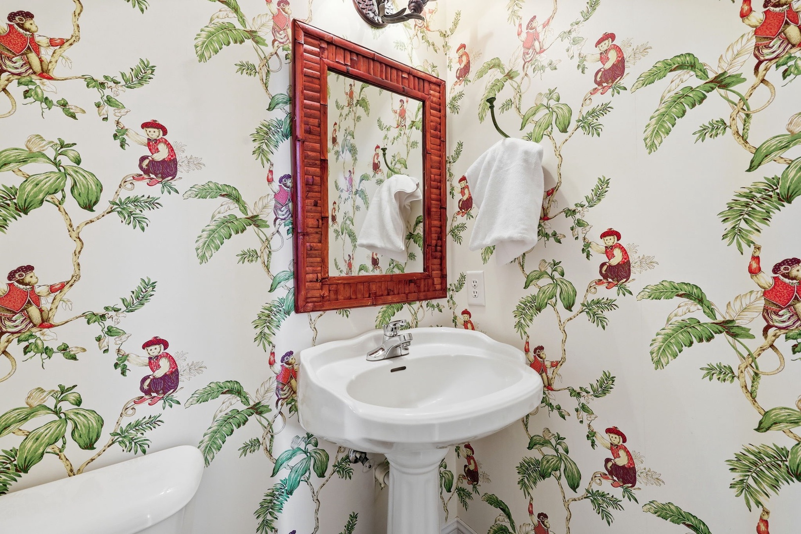Freshen up in this whimsical bathroom adorned with playful tropical wallpaper. The classic pedestal sink and bamboo-framed mirror add a charming, cottage-style touch to your stay.