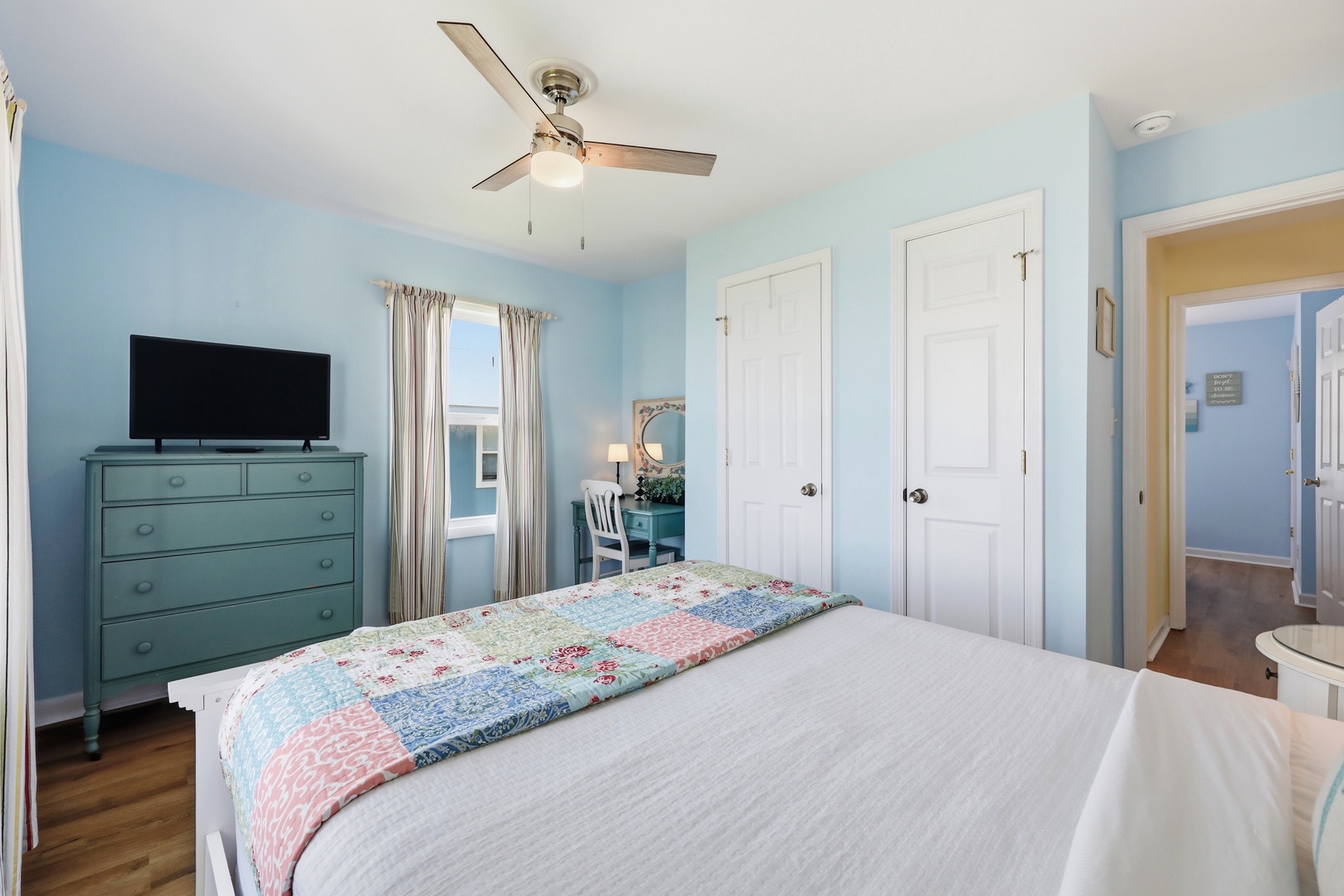 Picture yourself waking up in this serene coastal-inspired bedroom. Soft blue walls and a gentle ceiling fan create a refreshing retreat, while your flatscreen TV awaits your favorite shows after exploring.
