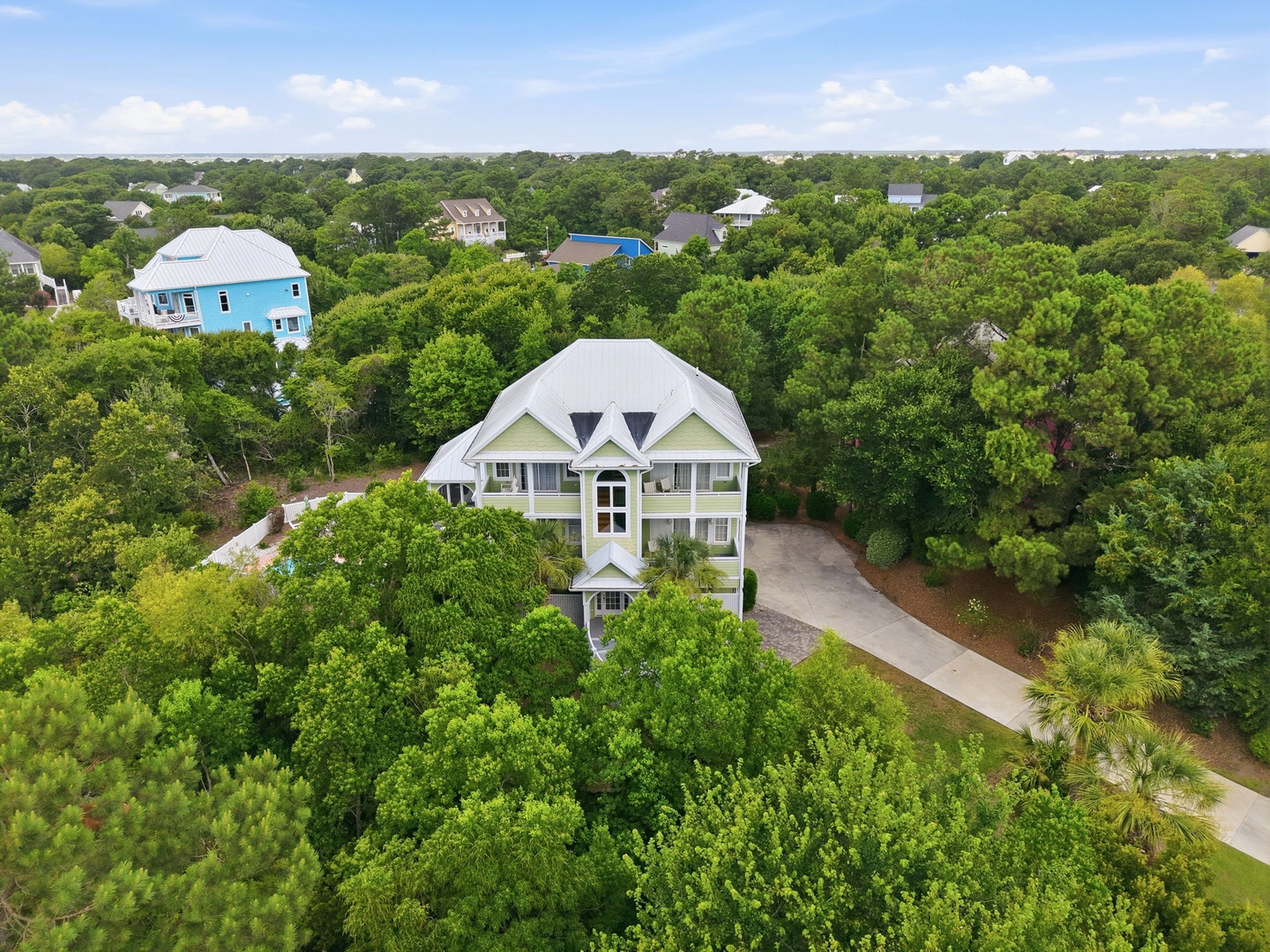 Aerial view showcases a spacious three-story coastal home with a metal roof, nestled among lush oak trees in a quiet residential neighborhood, with convenient parking and nearby community amenities.