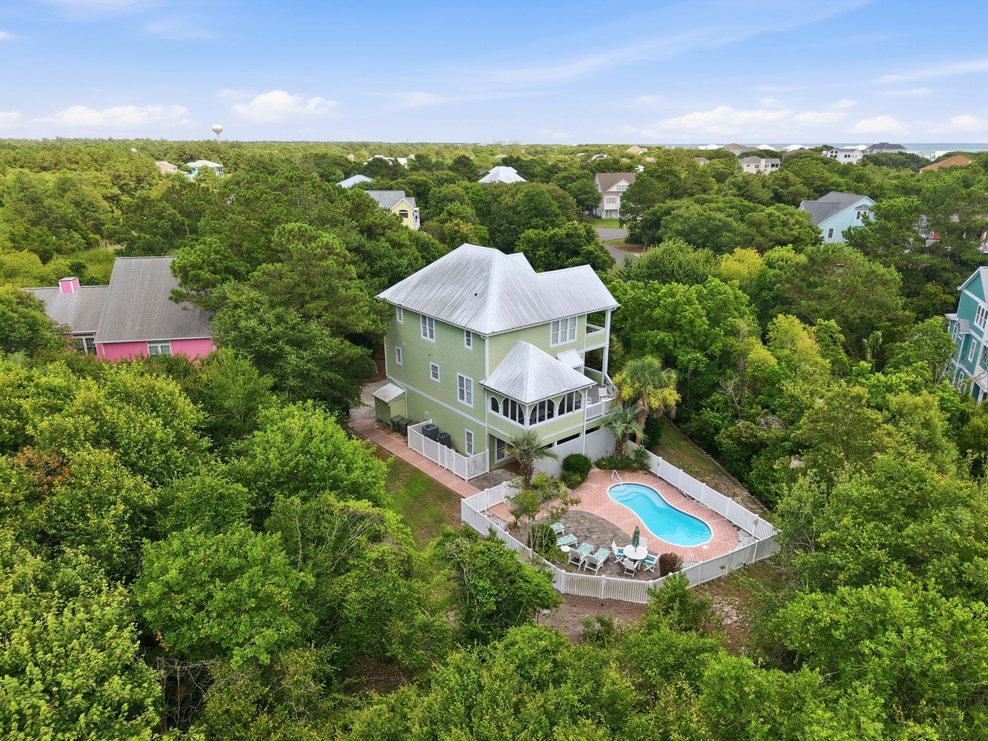 Aerial view of a spacious green vacation home nestled among coastal trees, featuring a private pool surrounded by lounge chairs, white fencing, and tiered decking in a quiet residential neighborhood.