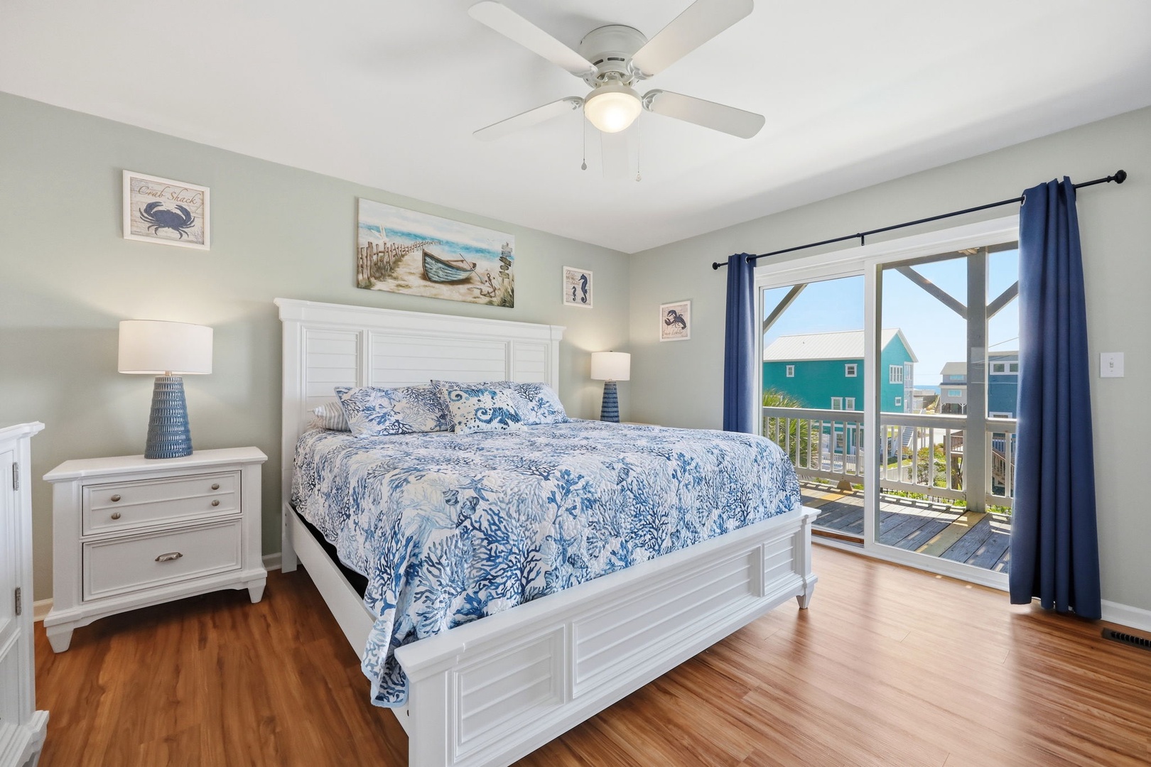 Wake up to bright coastal views and ocean breezes in this serene bedroom. Plush bedding, warm wood floors, and a private balcony overlooking colorful beach homes make mornings here unforgettable.