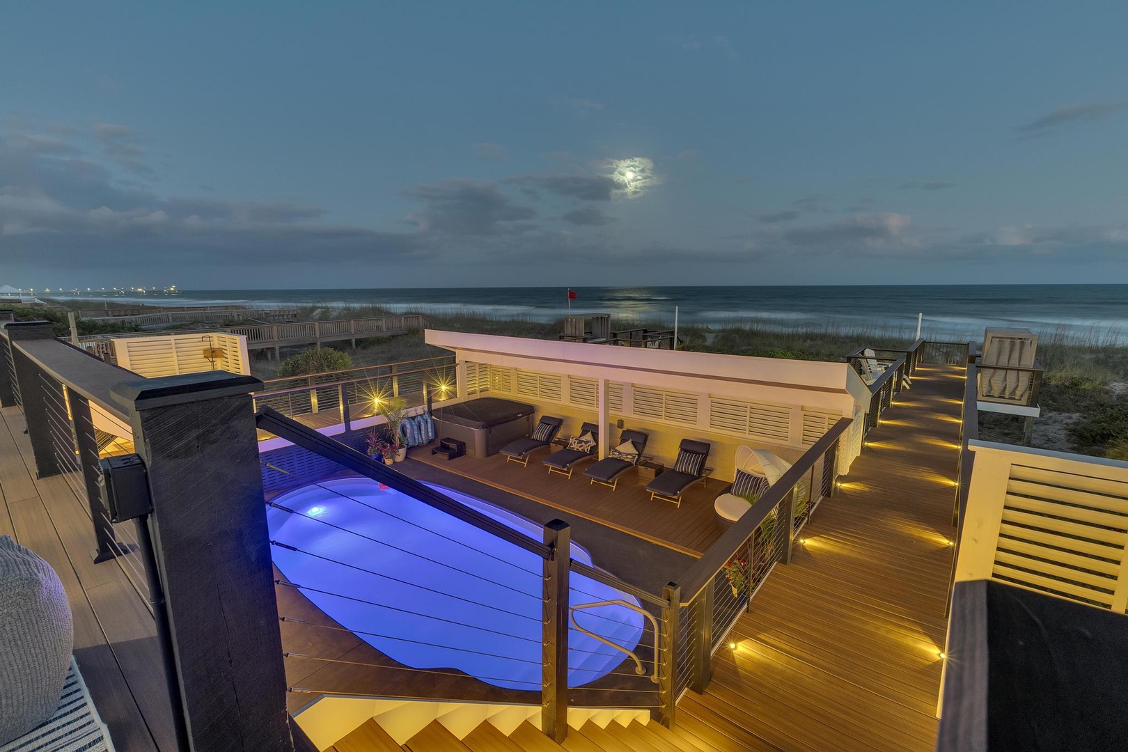 Elevated beachfront deck with illuminated pool, ocean views, and modern outdoor amenities under the evening sky.