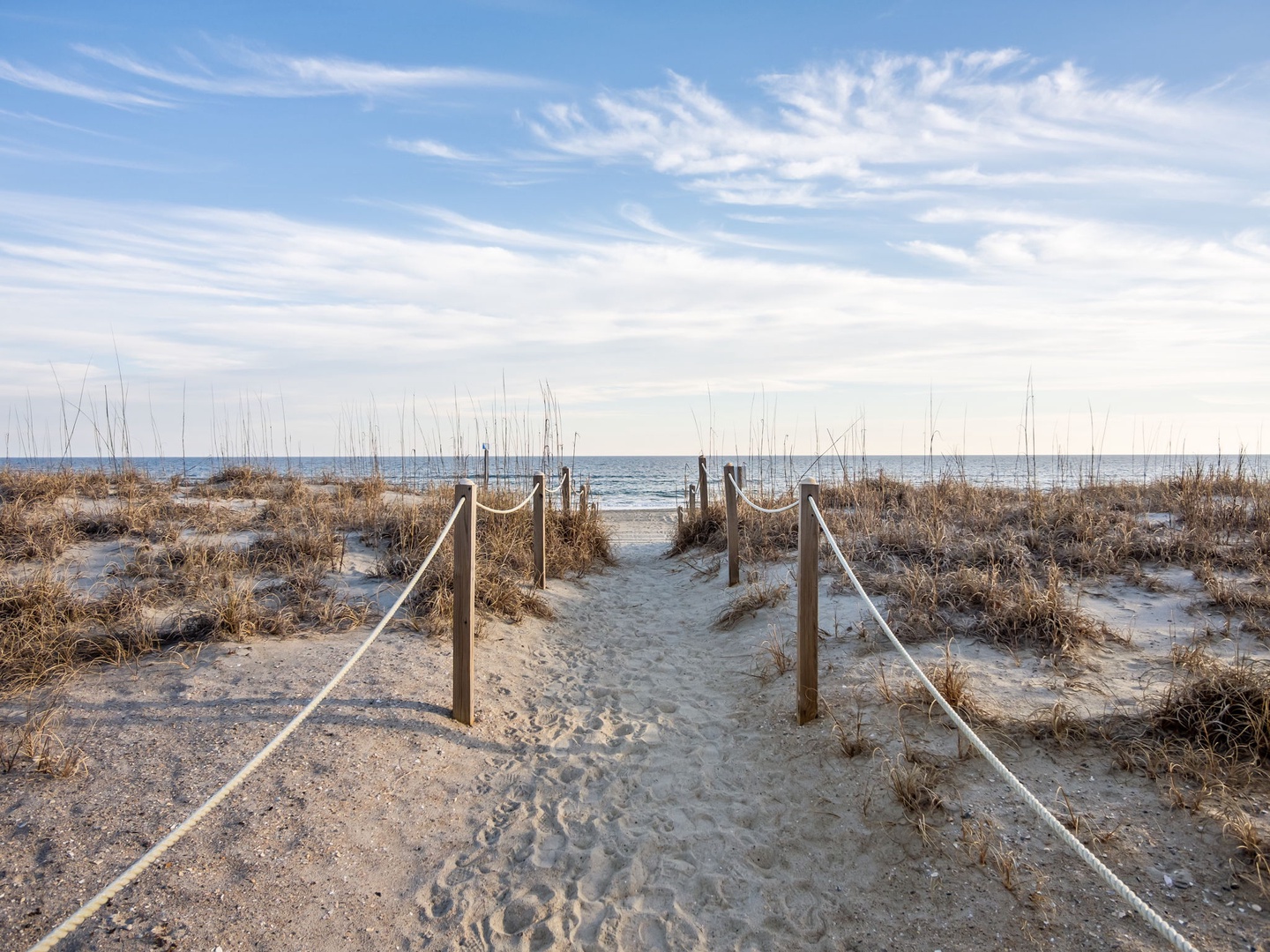 Sandy beach pathway with rope railings leads through dunes to the ocean shoreline.