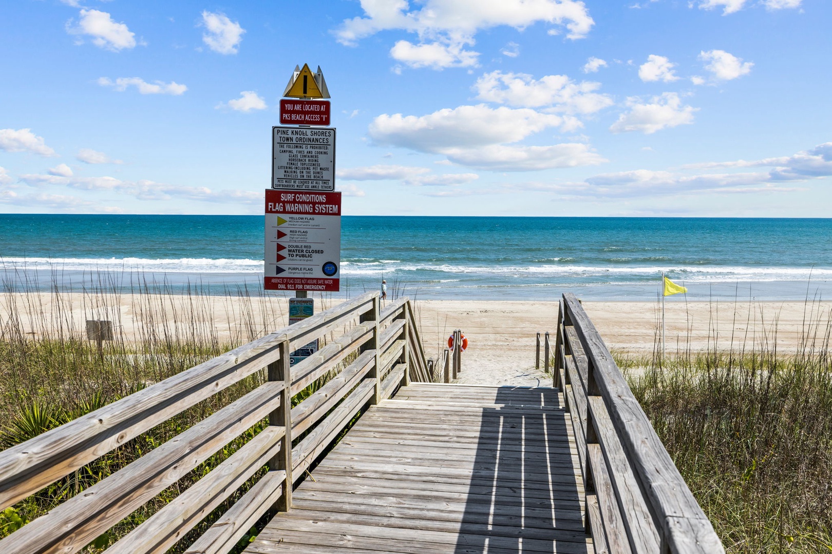 Wooden boardwalk leads to pristine beach with clear blue waters and gentle waves under sunny skies.