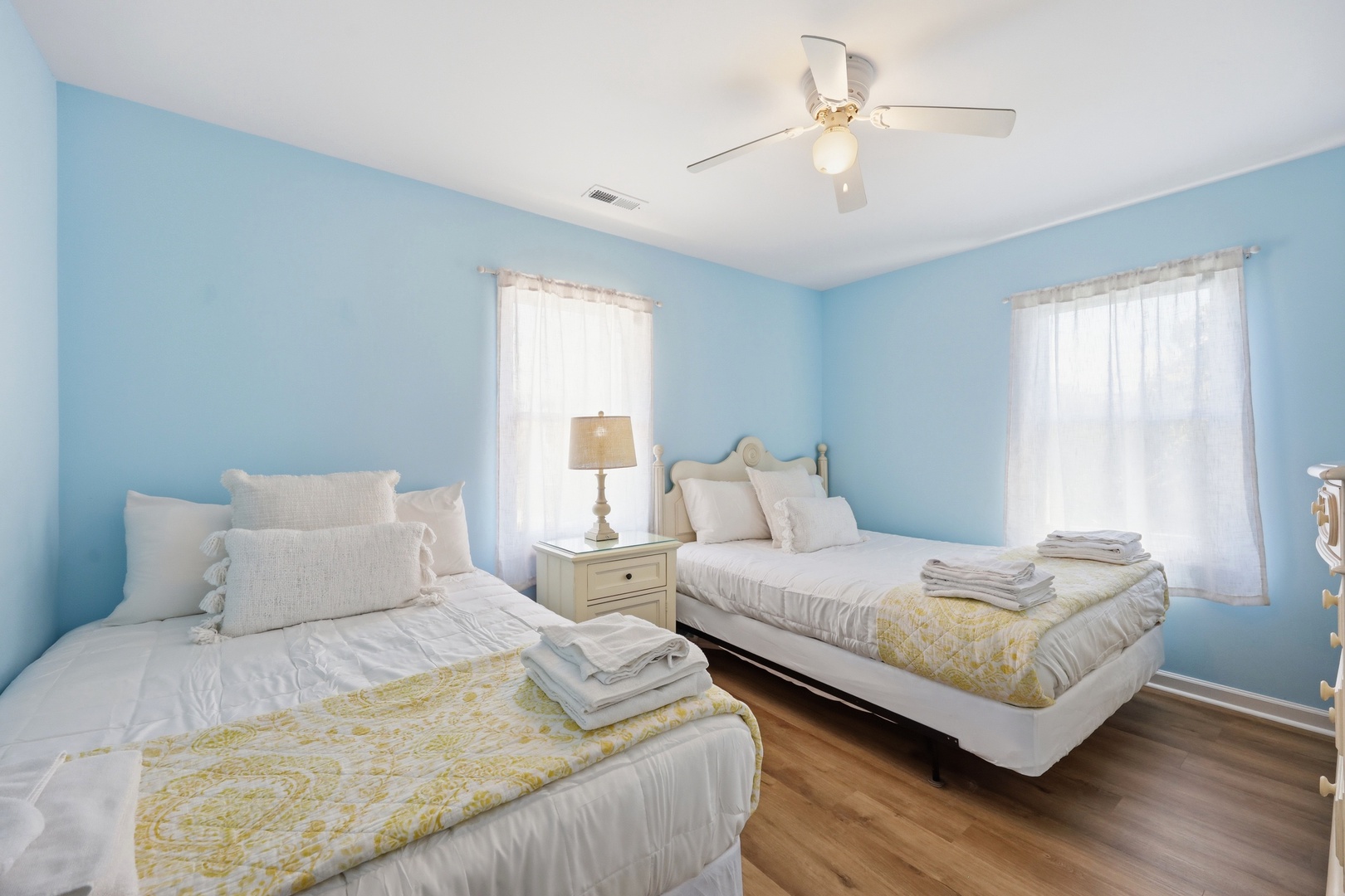 Drift off in this tranquil bedroom with two cozy beds, soft blue walls, natural light, and a gentle ceiling fan overhead keeping you comfortable.