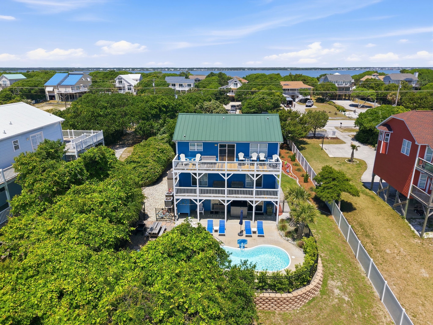 Aerial view of a blue waterfront vacation home featuring a private pool and spacious decks, situated in a coastal neighborhood with lake views beyond.