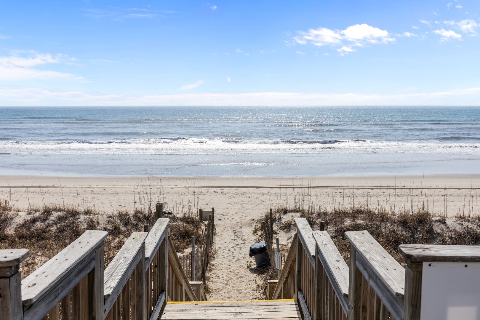 Wooden boardwalk leads directly to pristine sandy beach with gentle waves rolling onto shore under bright blue skies.