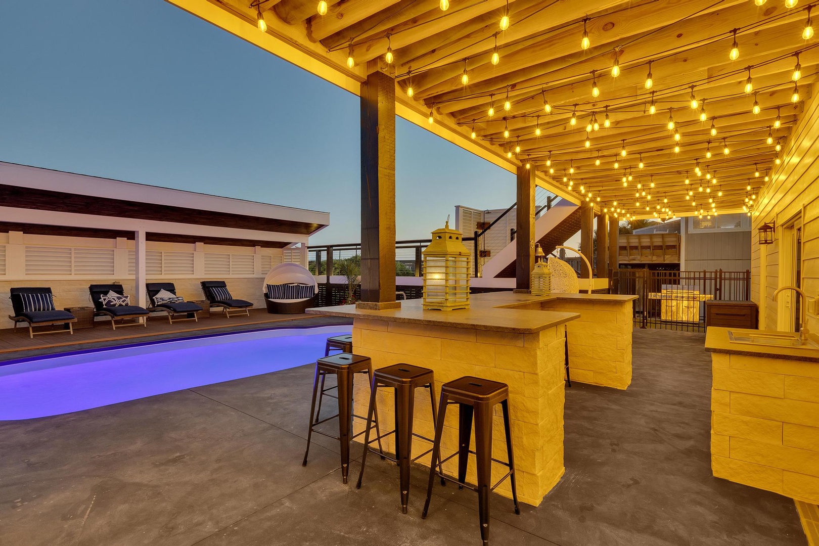 Stunning rooftop lounge area with illuminated pergola, poolside bar seating, and modern outdoor entertaining space as evening sets in.