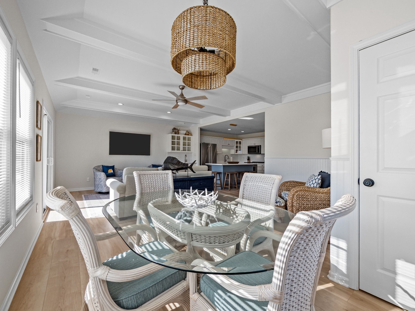Your bright open-concept living space blends coastal charm with modern comfort, featuring wicker dining chairs and flowing natural light.