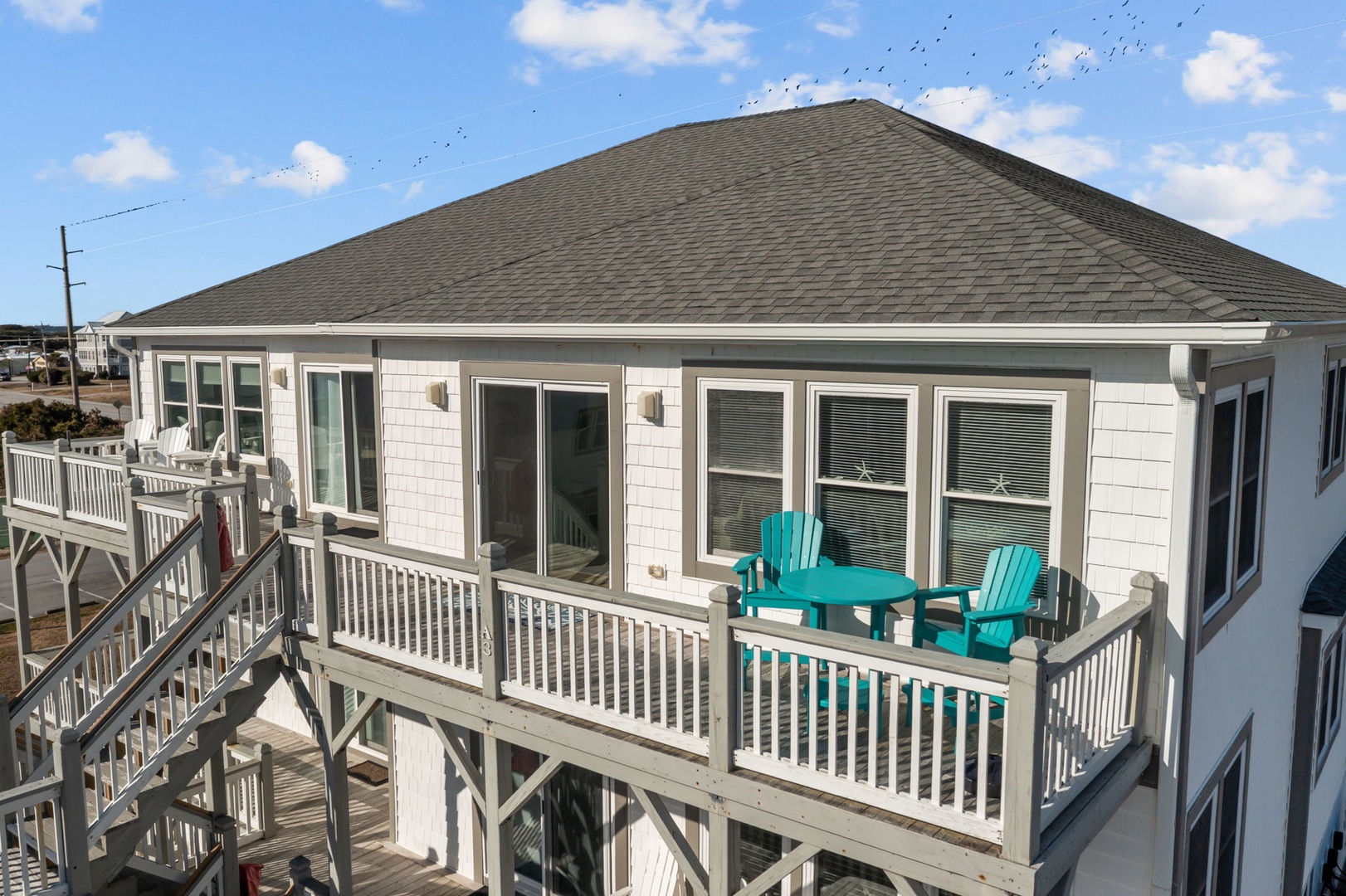 Coastal rental property featuring elevated deck with turquoise chairs and white railings under blue skies.