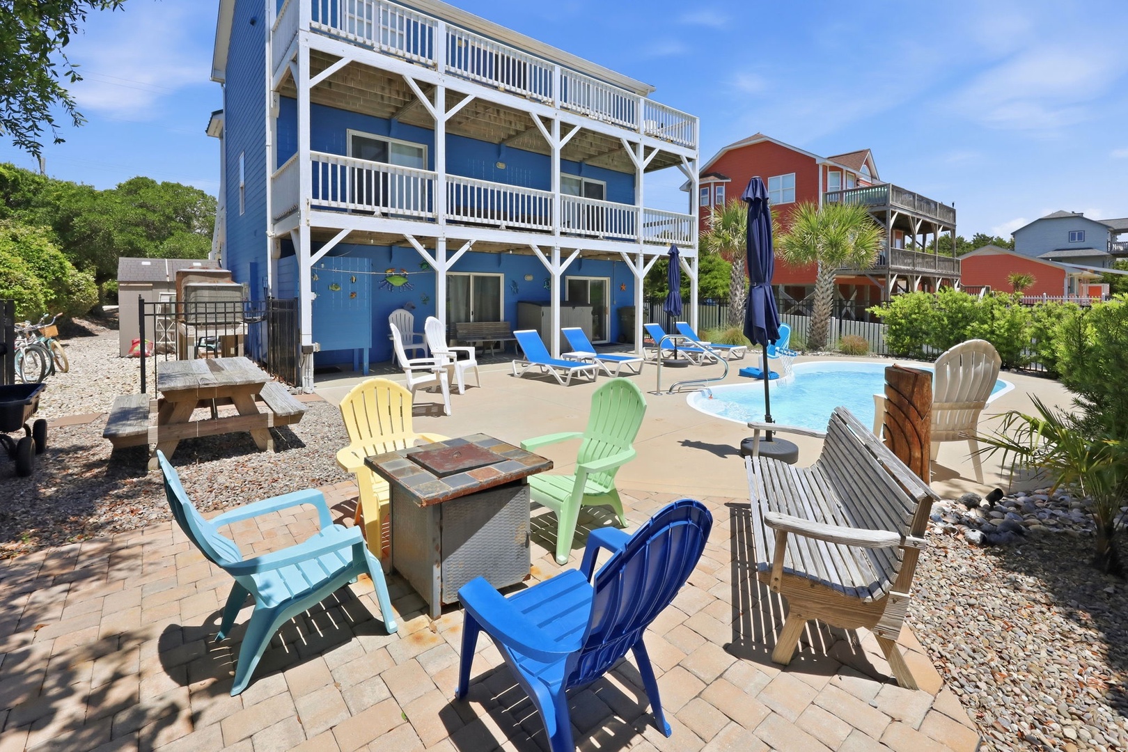 Bright coastal retreat with private pool, sun loungers, colorful Adirondack seating around a fire pit, grilling area, outdoor dining, and shaded lounging—perfect for lazy beach days.