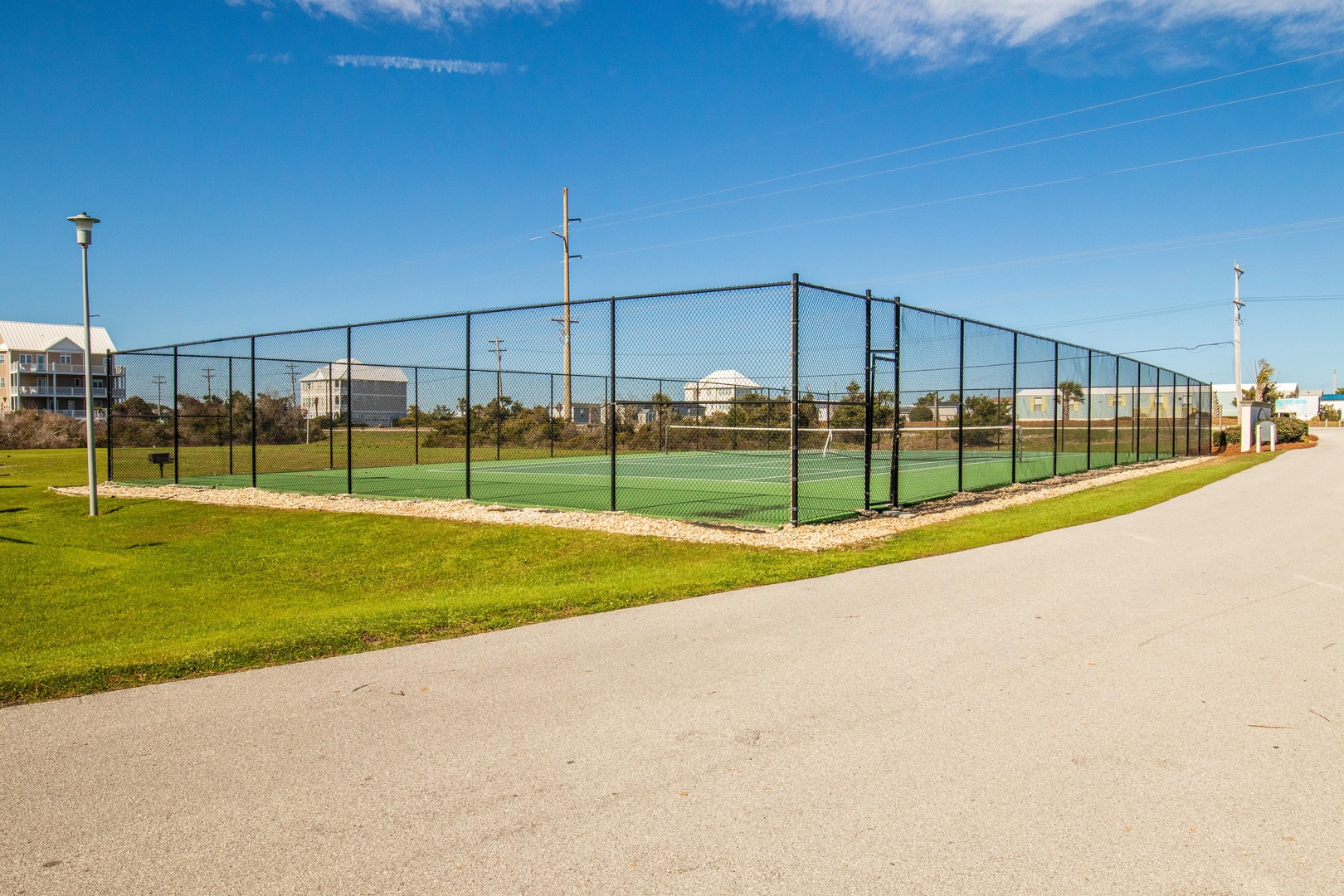 Tennis court with chain-link fencing surrounded by green lawn and residential buildings in the background.