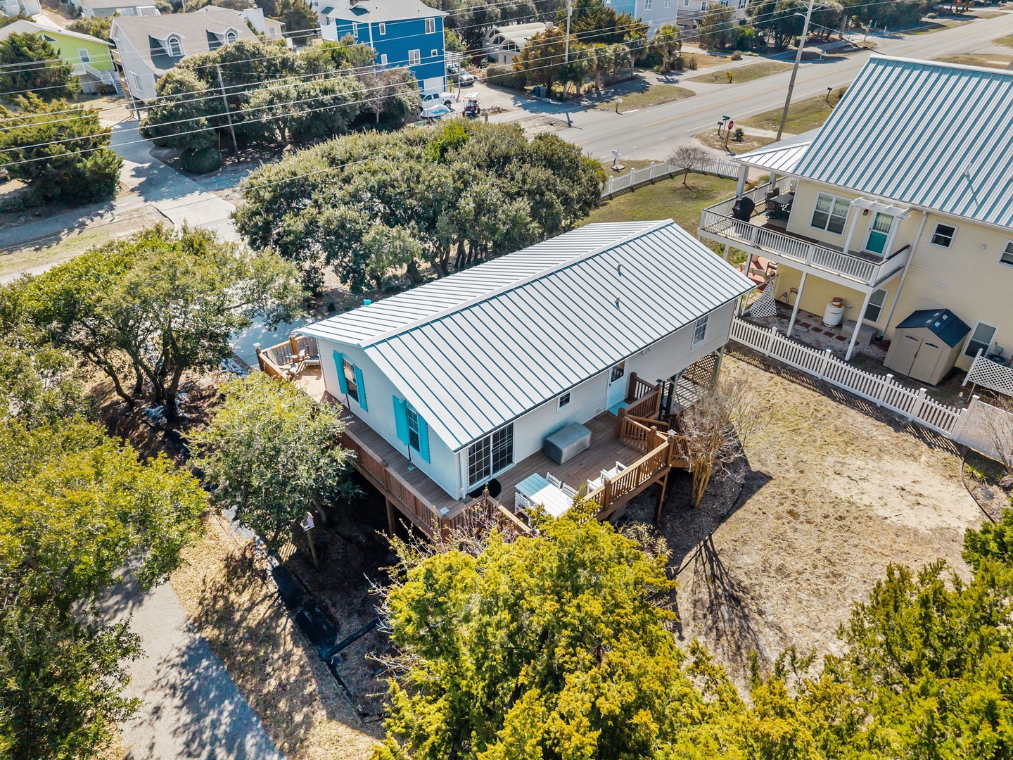 Aerial view of a charming coastal vacation home with metal roof and wraparound deck, nestled among mature trees in a quiet residential neighborhood.