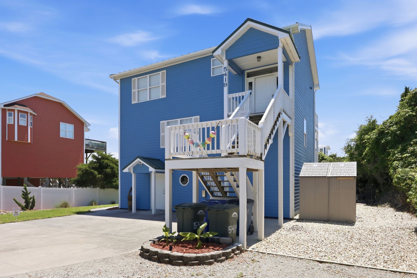 Bright blue coastal beach house with elevated deck and covered parking, located in a quiet residential neighborhood near the shore.