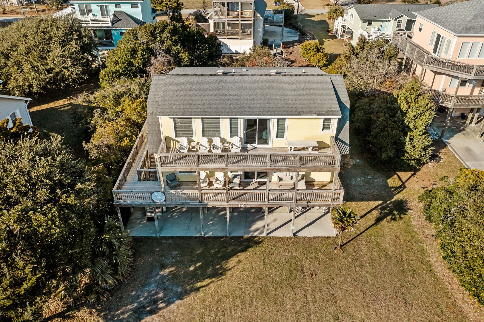 Aerial view of a charming coastal rental property with wraparound decks and outdoor seating, nestled among neighboring beach homes.