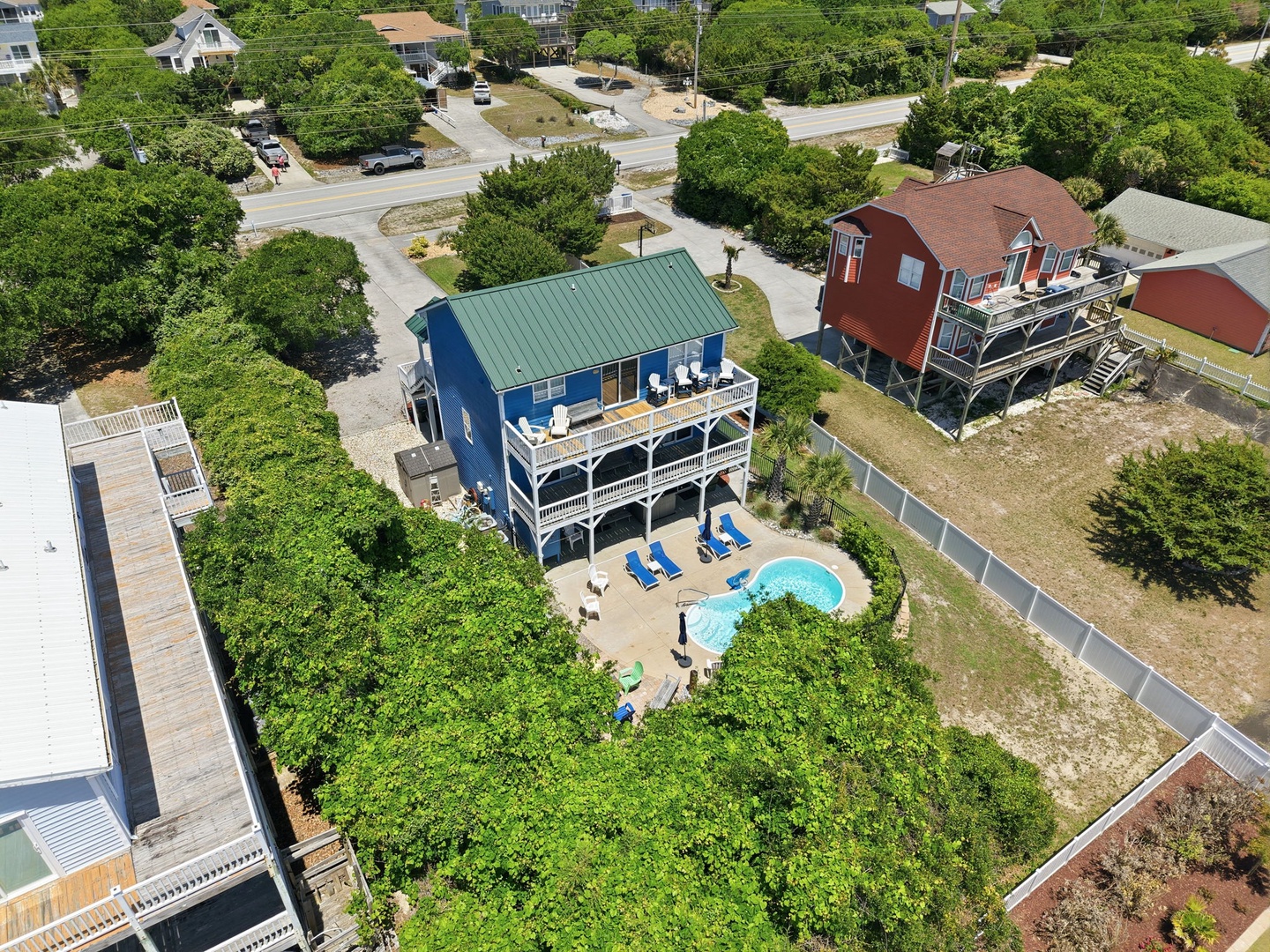 Aerial view of a charming blue coastal vacation rental nestled among lush greenery, featuring a private pool and spacious deck in a peaceful neighborhood setting.