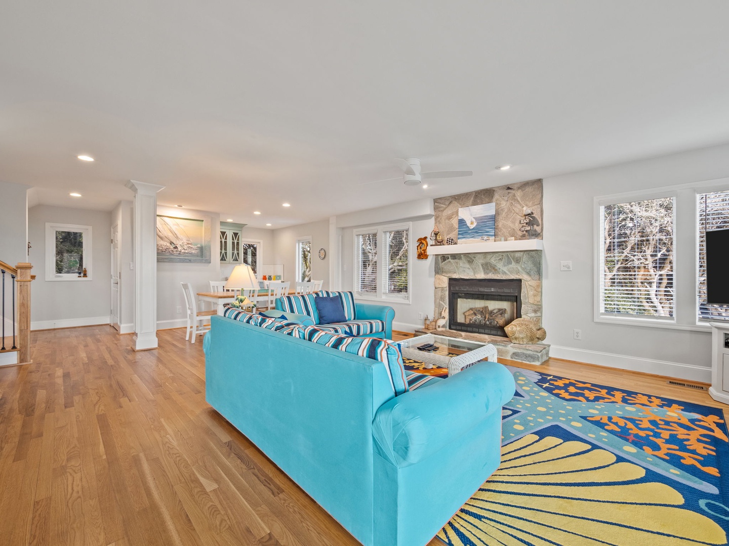 Relax in your bright, coastal-inspired living room with vibrant blue sectional seating, crackling stone fireplace, and panoramic windows overlooking the trees.