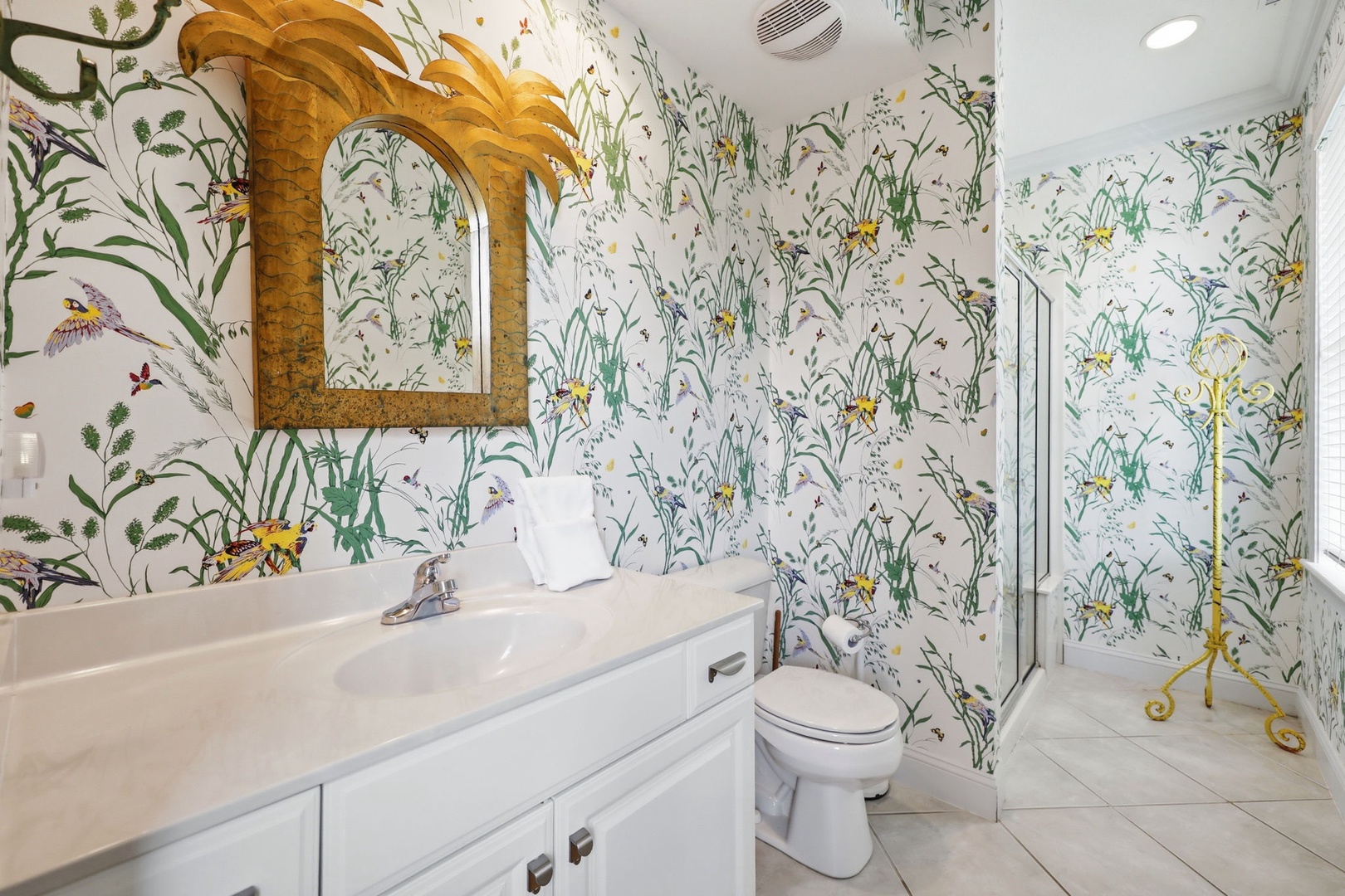 A vibrant botanical-themed bathroom featuring whimsical wallpaper with birds and florals, a golden palm mirror, and a cheerful atmosphere that makes freshening up feel like a tropical escape.