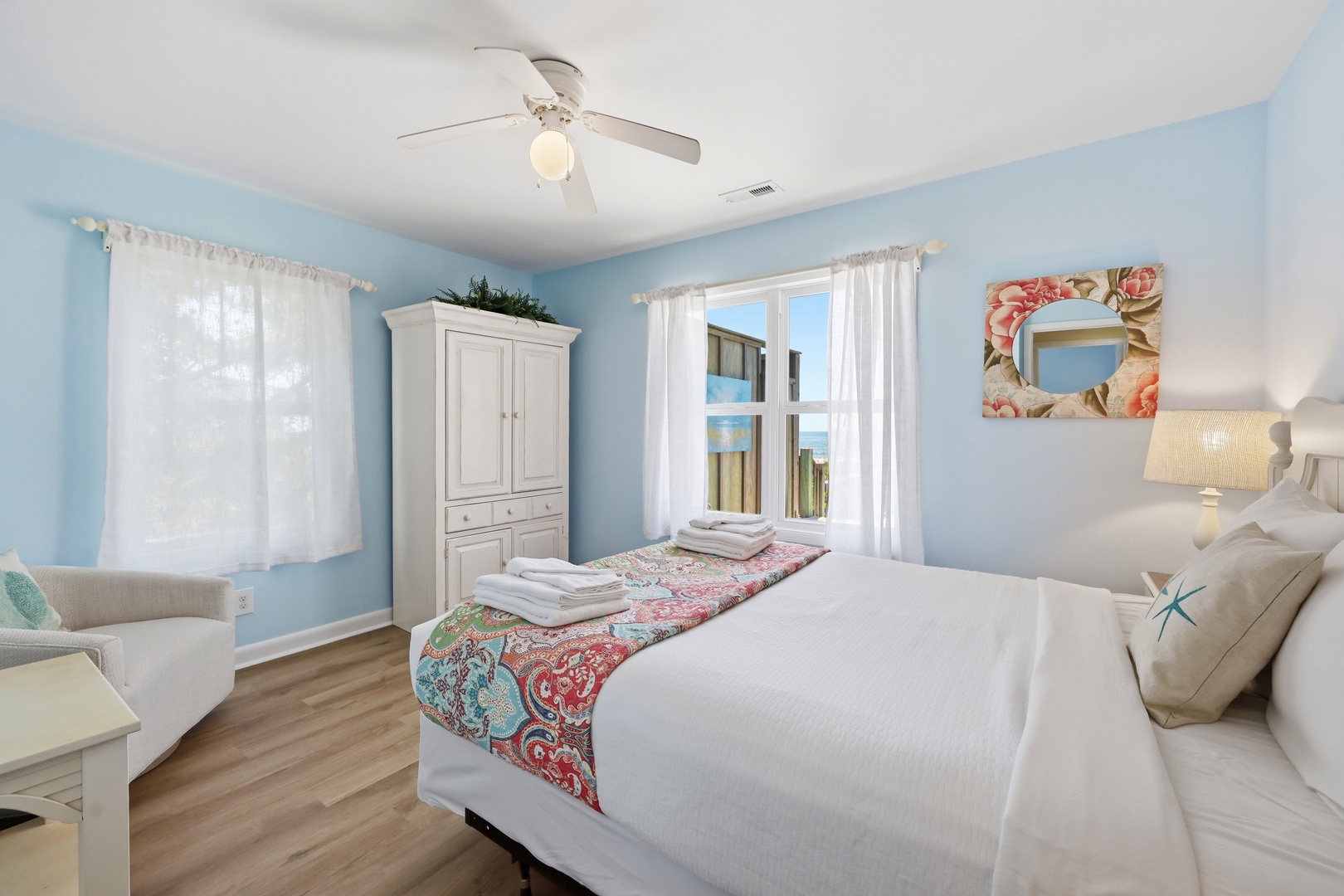 Your serene coastal bedroom features soothing blue walls, crisp white linens, and a ceiling fan to keep you cool. Natural light fills the space, while beach-inspired decor invites relaxation.