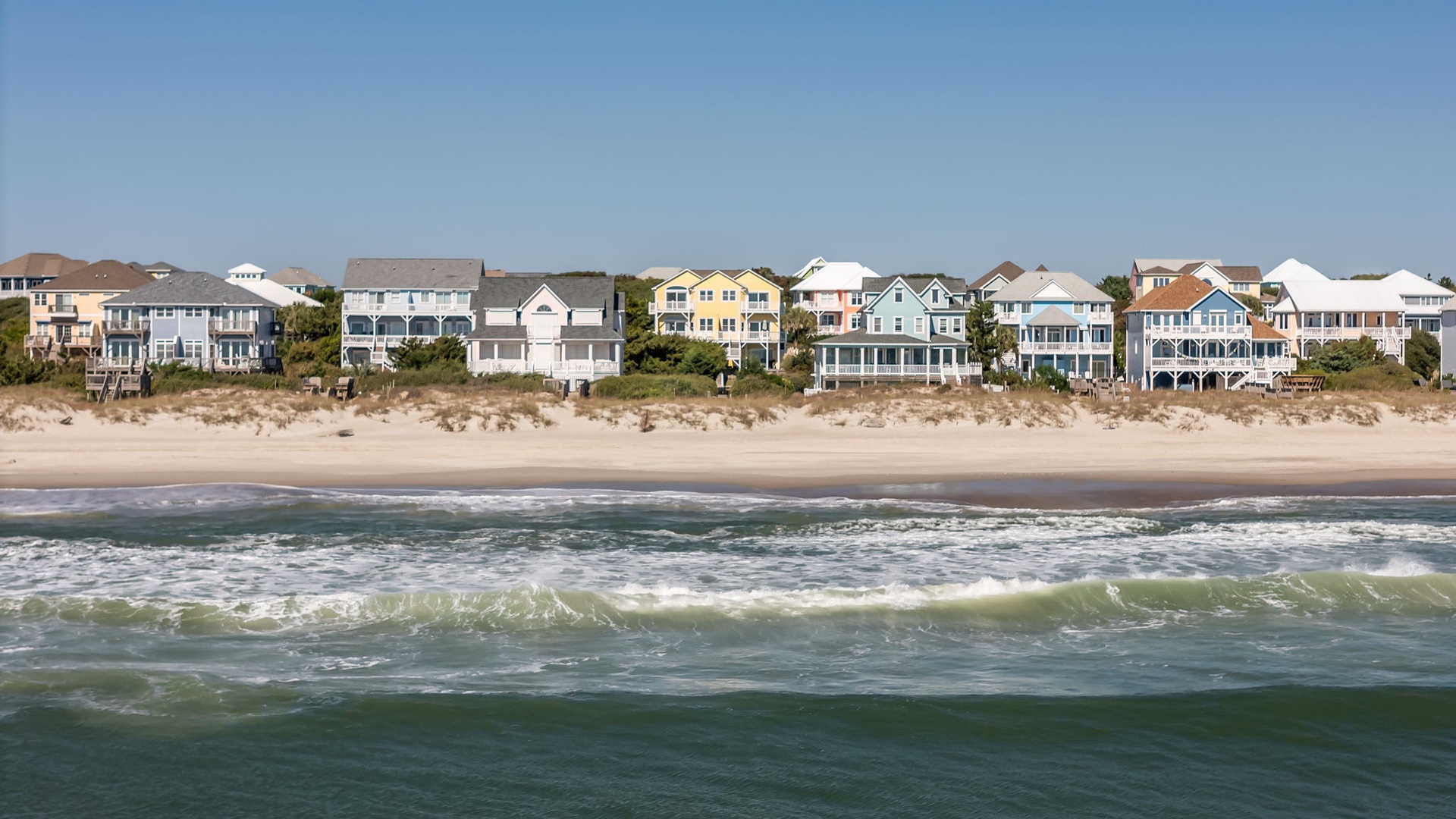Beachfront vacation homes line a pristine sandy shore, with ocean waves rolling in under clear blue skies. A serene coastal setting perfect for seaside getaways.