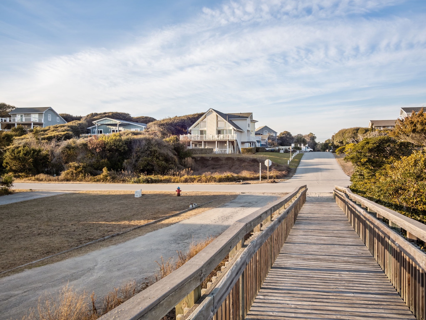 Wooden boardwalk leads through coastal neighborhood with beach houses nestled among dunes and native vegetation.