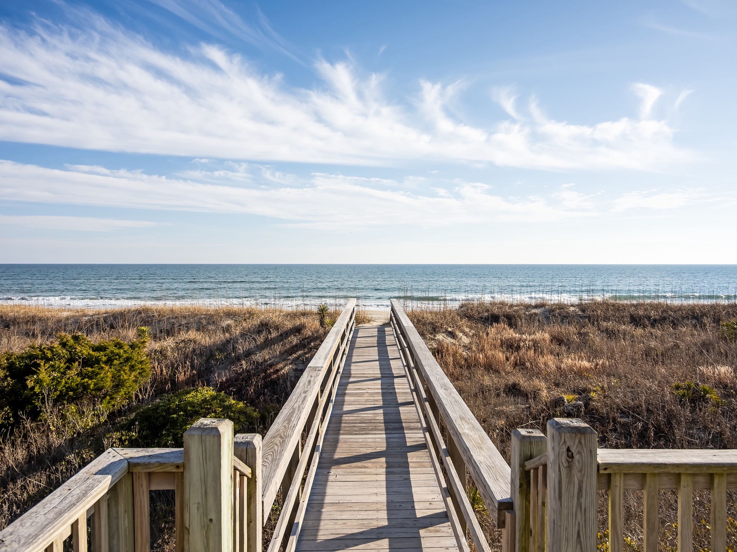 A wooden boardwalk leads through coastal dunes to pristine beach access under expansive blue skies.