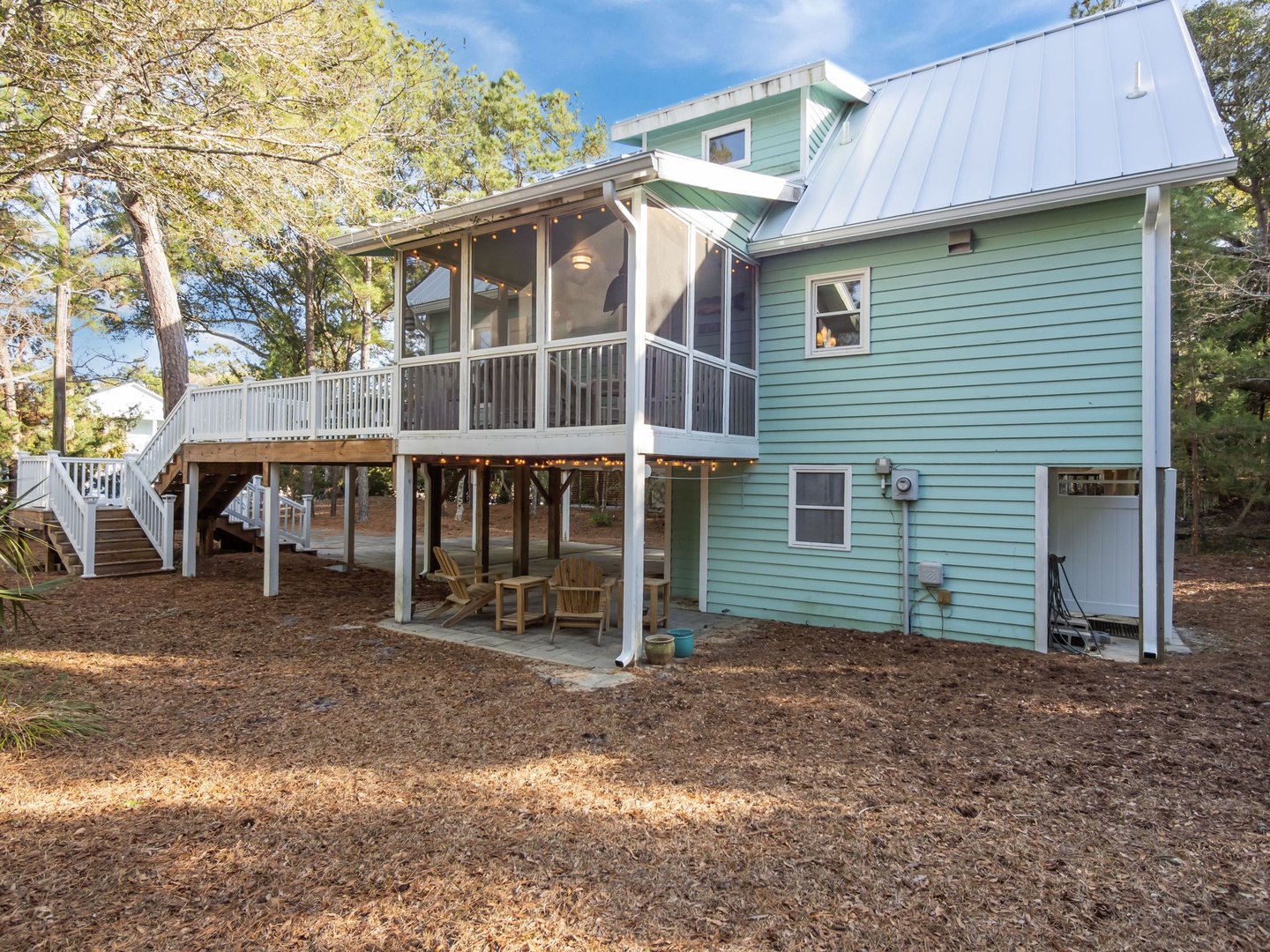 Charming coastal cottage featuring elevated deck with string lights and ground-level patio surrounded by mature trees.