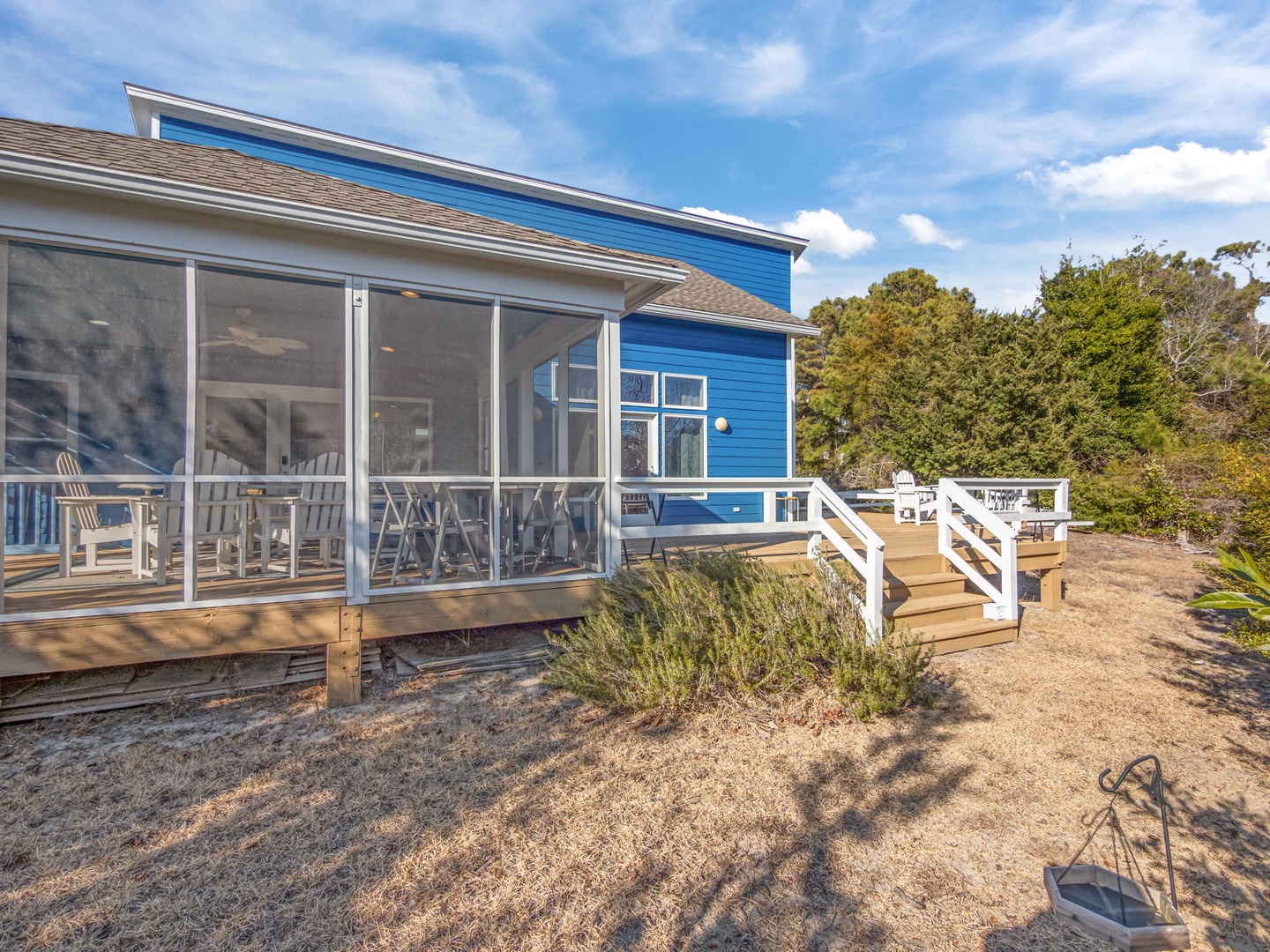 Charming blue coastal cottage with screened porch and deck surrounded by mature trees under bright blue skies.