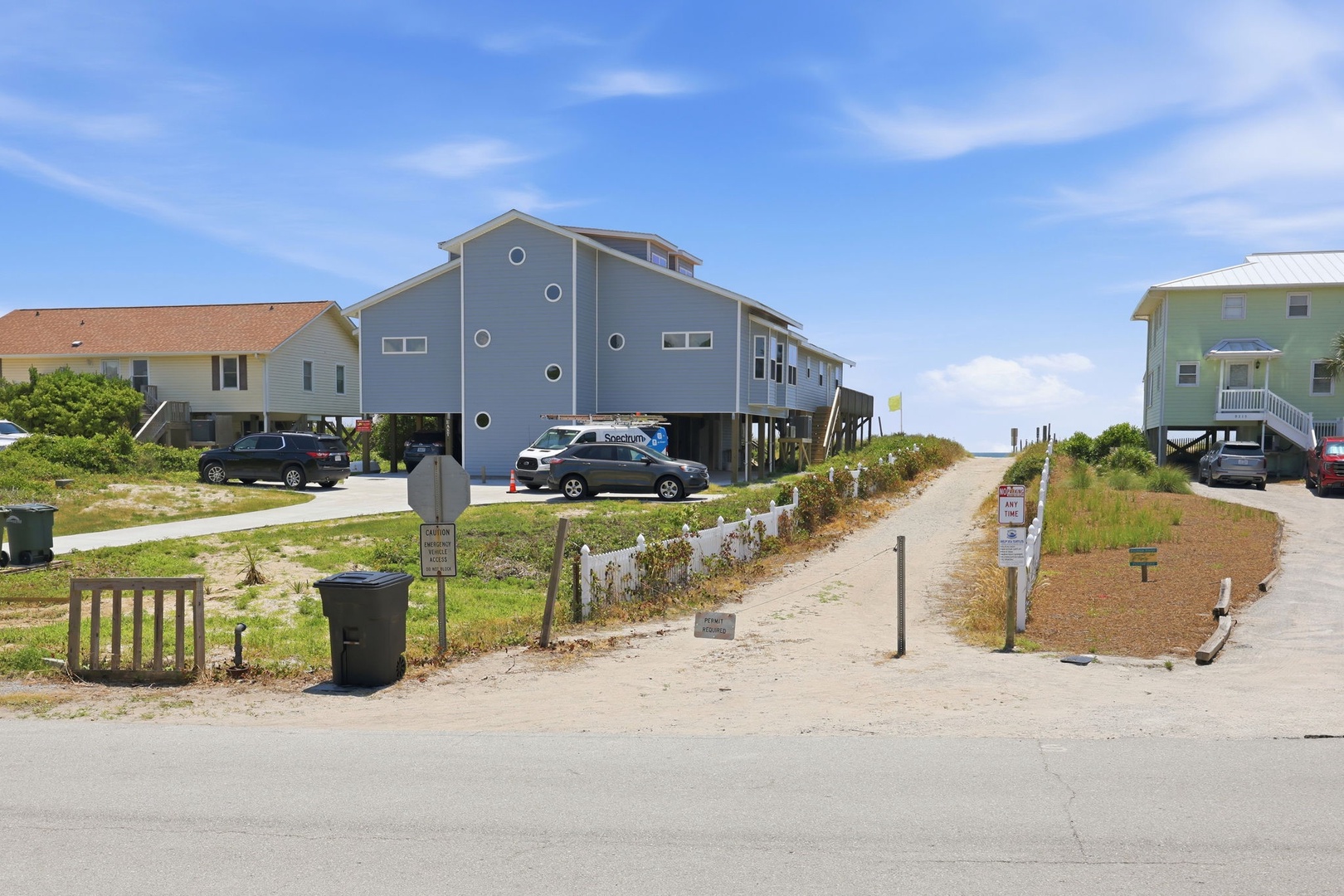 Coastal vacation homes elevated on stilts with direct beach access through a sandy pathway, surrounded by natural dune vegetation under clear blue skies.