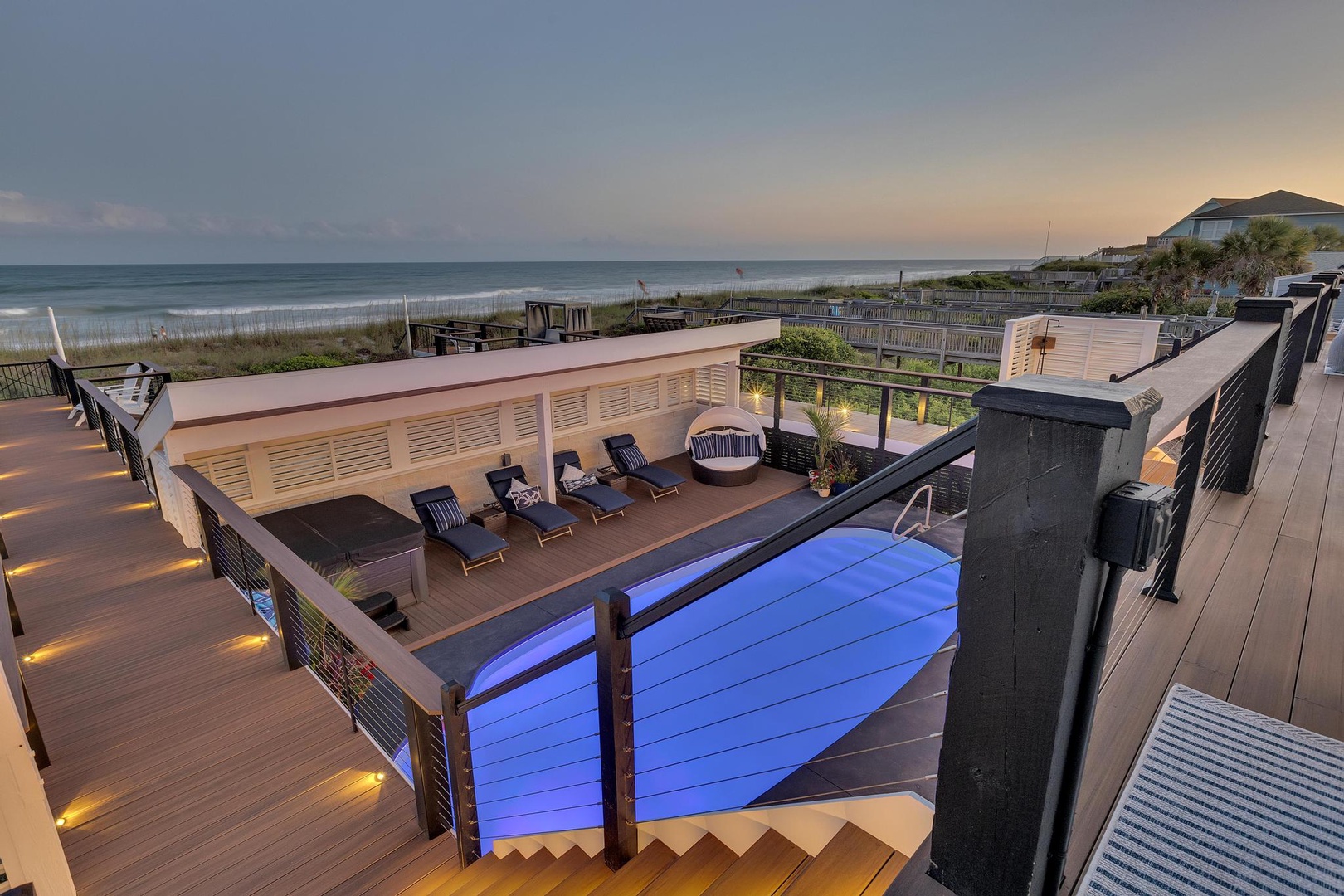 Oceanfront property with expansive deck featuring an illuminated pool, lounge seating, and direct beach access via wooden walkway.