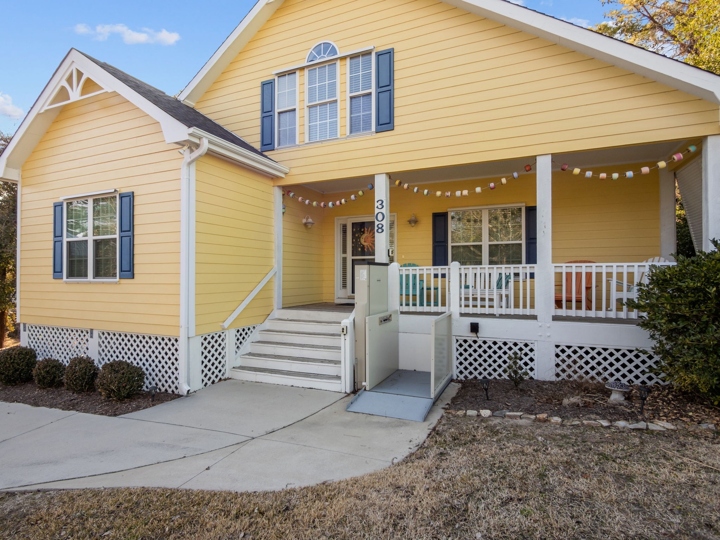 Charming yellow cottage with welcoming front porch and accessibility ramp, featuring festive string lights and comfortable outdoor seating area.