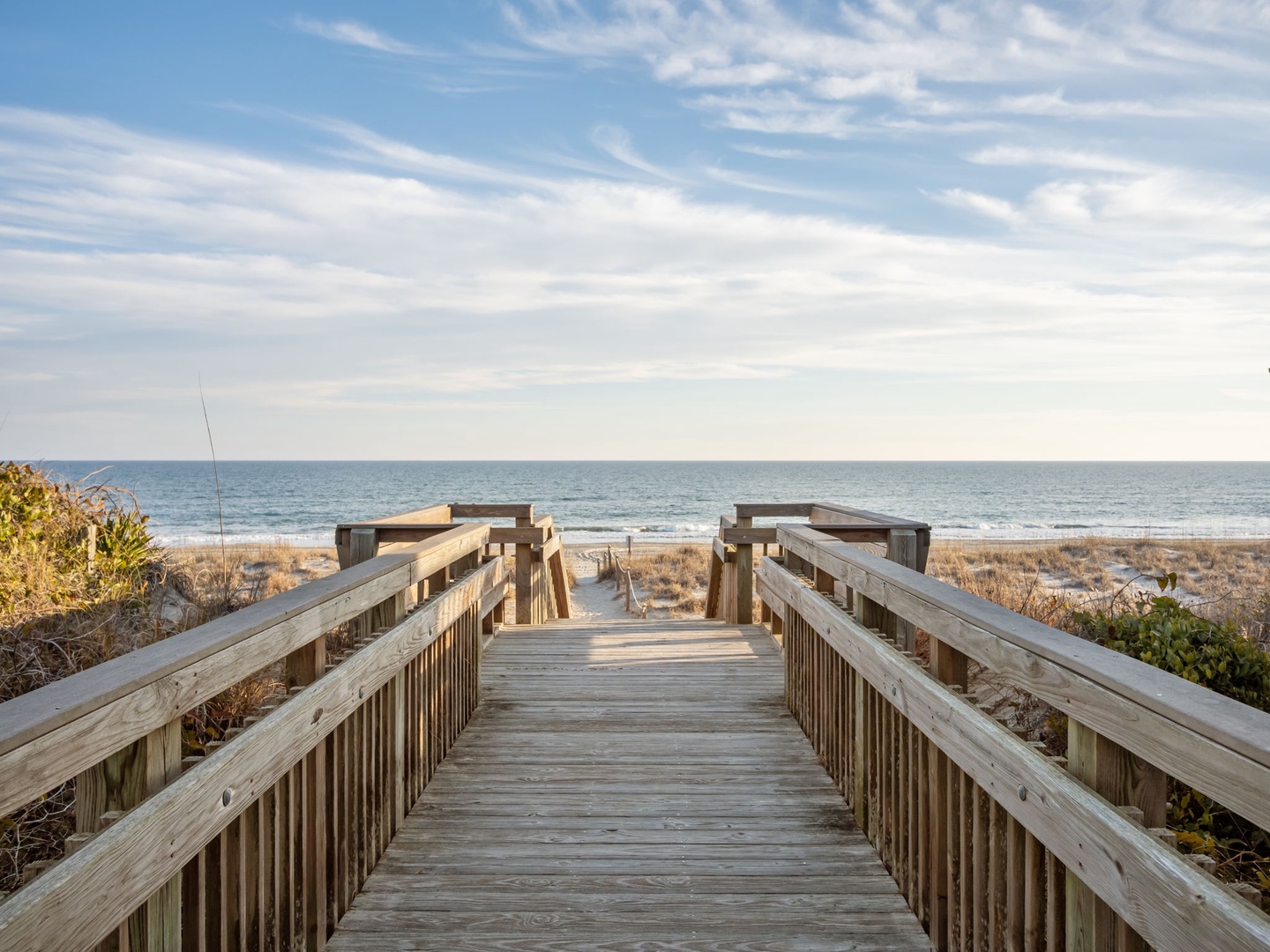 Wooden boardwalk leads to pristine beach with ocean waves and coastal dunes under expansive sky.