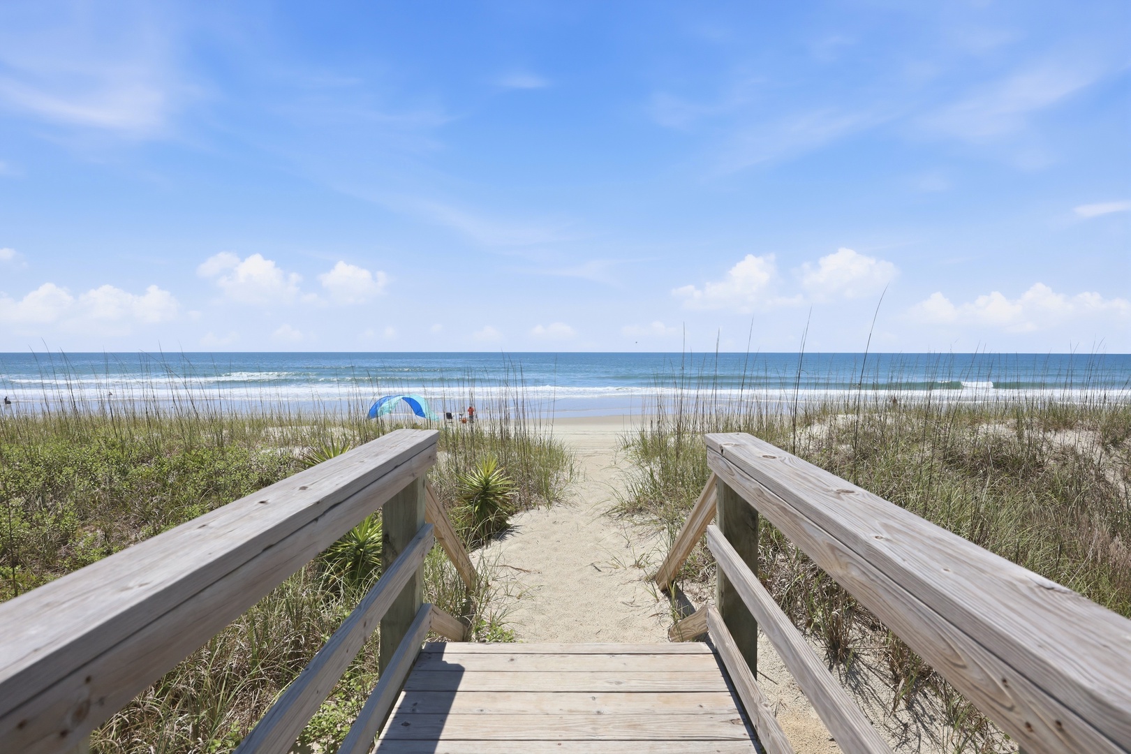 A private wooden boardwalk leads through gentle dunes, offering direct access to the wide sandy shore and sparkling ocean waves beyond.