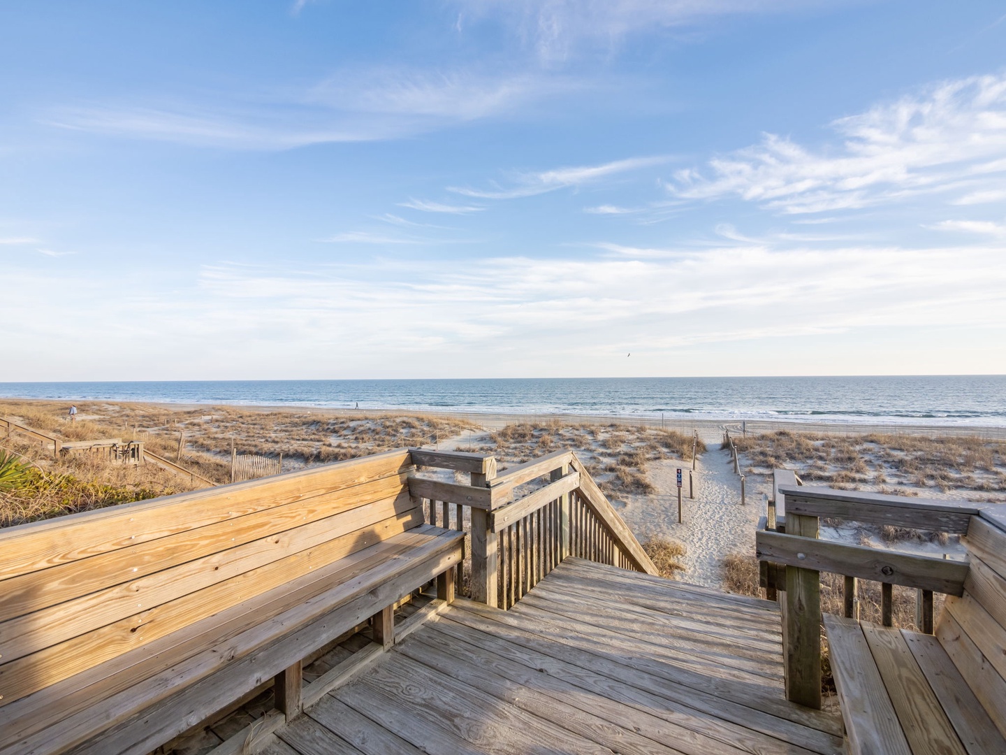 Wooden boardwalk leads through coastal dunes to pristine oceanfront beach under expansive sky.
