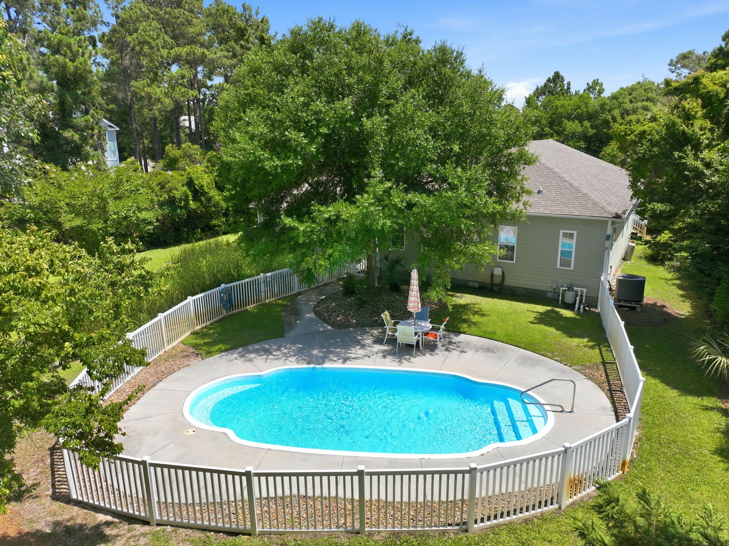Aerial view showcasing a private backyard oasis with kidney-shaped pool, surrounded by lush greenery and mature trees. The property features a secure white fence, spacious lawn, and shaded patio seating.