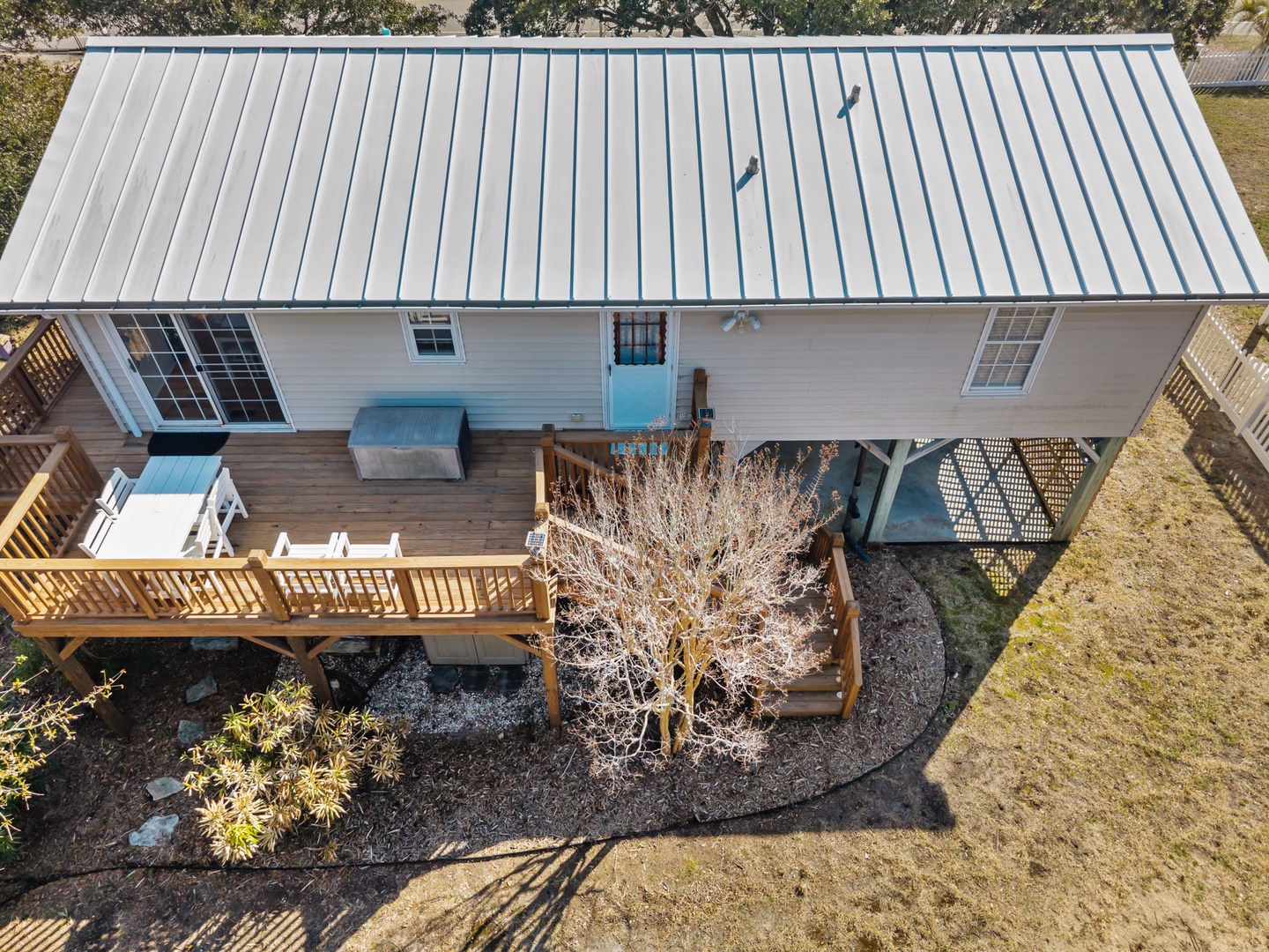 Charming beach house with metal roofing and wraparound deck, surrounded by natural landscaping and mature trees.