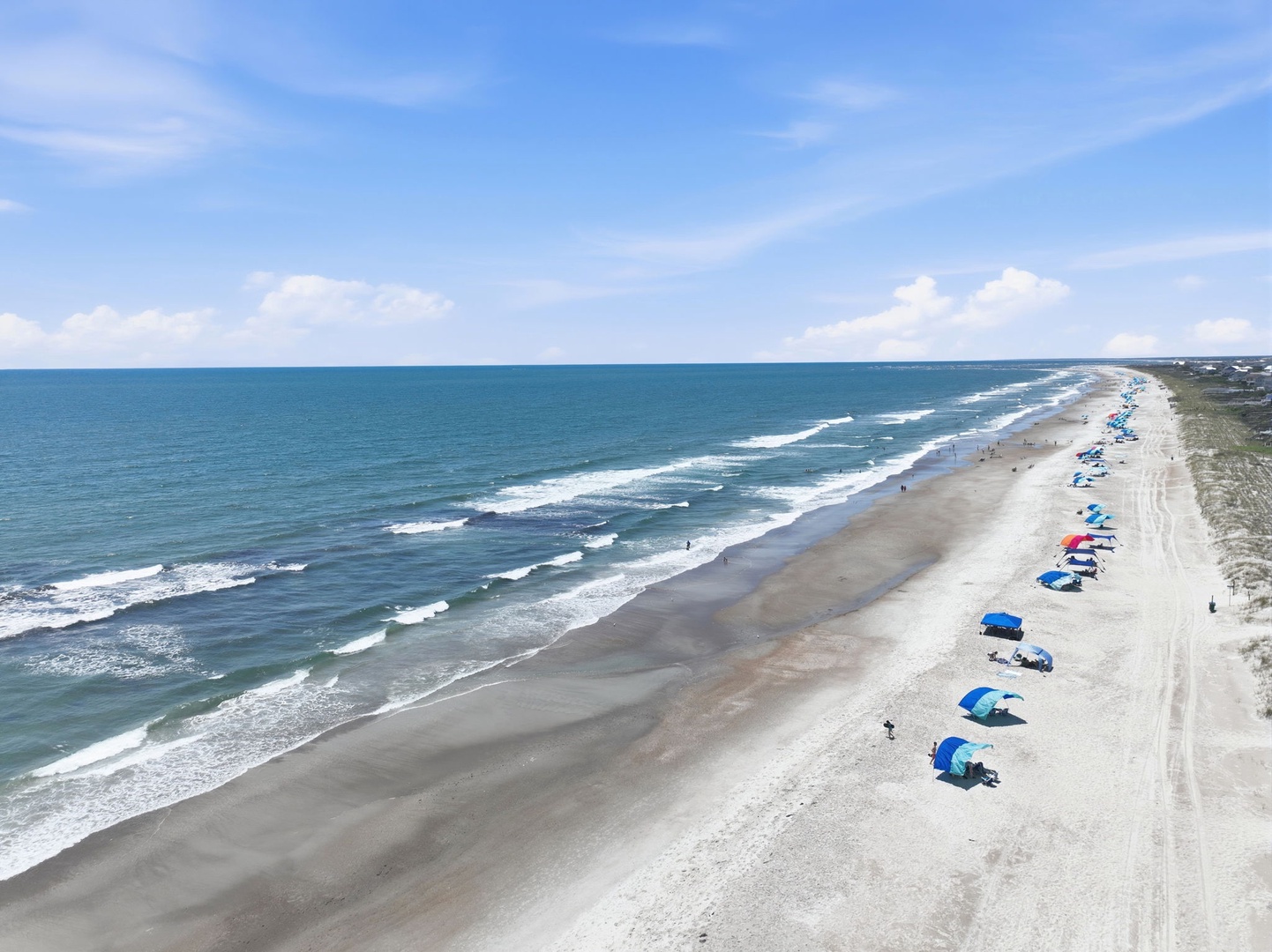 An aerial view captures a pristine stretch of sandy beach along the Atlantic coastline, with colorful umbrellas dotting the shore and gentle waves rolling in under a blue sky.