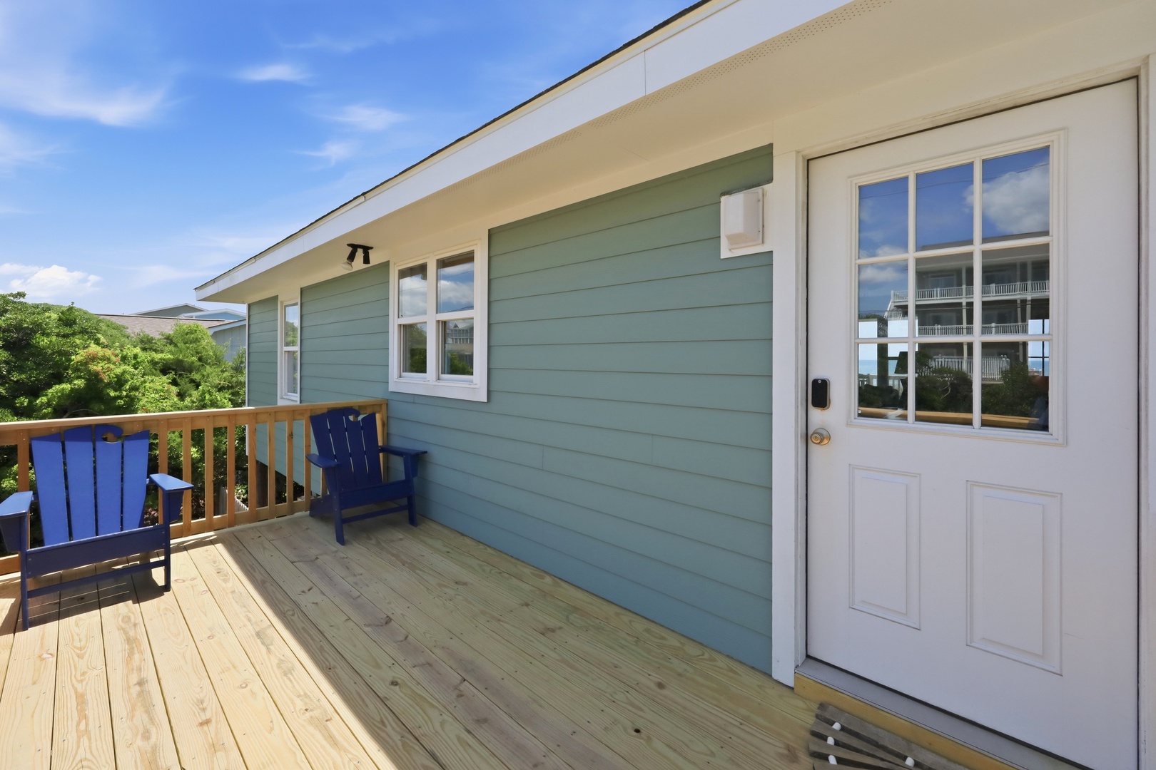 A welcoming coastal-style deck with blue Adirondack chairs and glass-paneled entrance door, offering a bright outdoor retreat with natural surroundings.