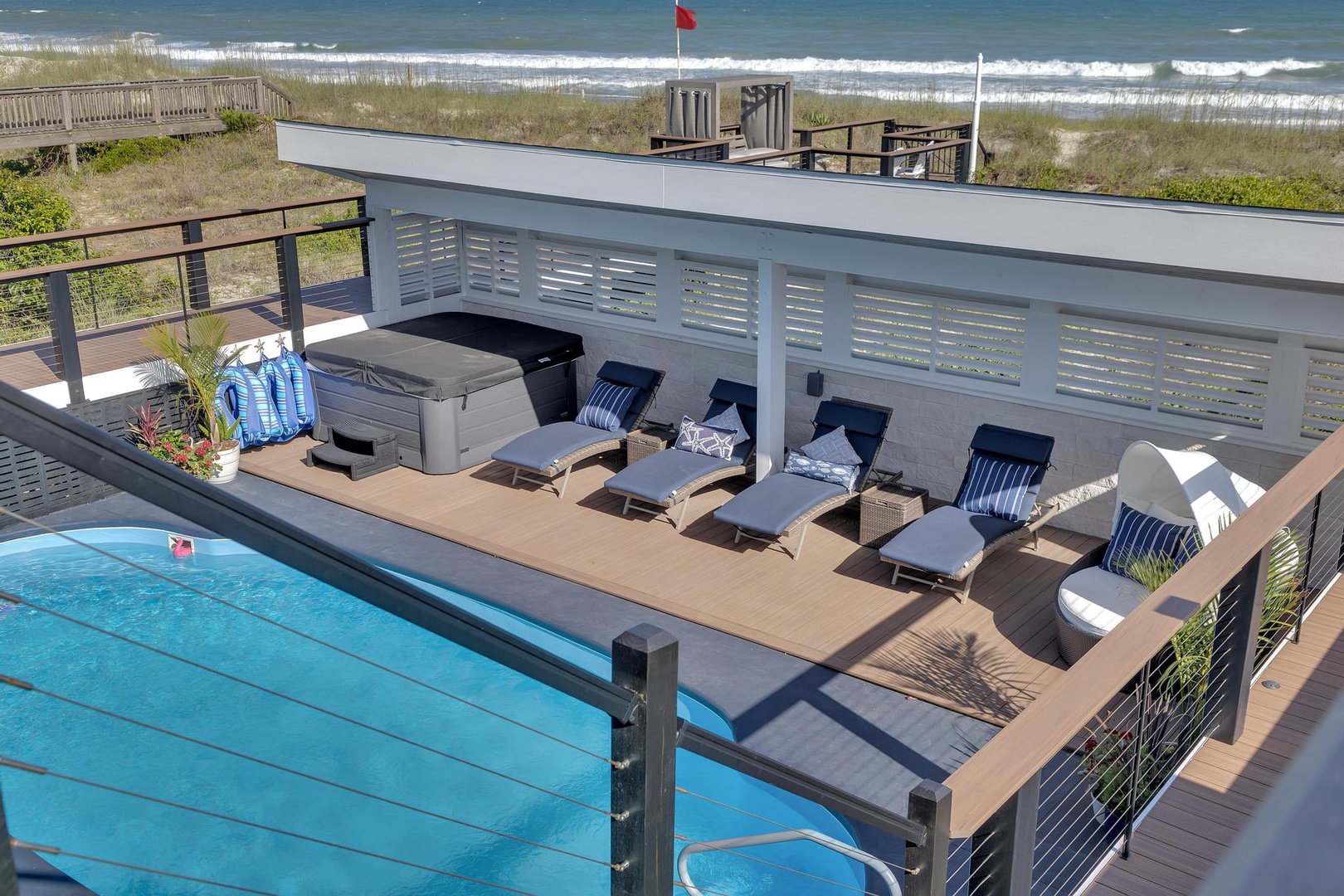 A coastal beachfront property features a rooftop pool deck with oceanfront access, surrounded by loungers, seating areas, and unobstructed views of the shore and rolling waves beyond.