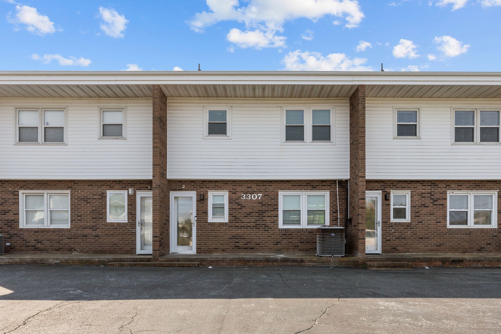 Multi-unit brick and vinyl siding building with individual entrances and parking area under bright blue skies.