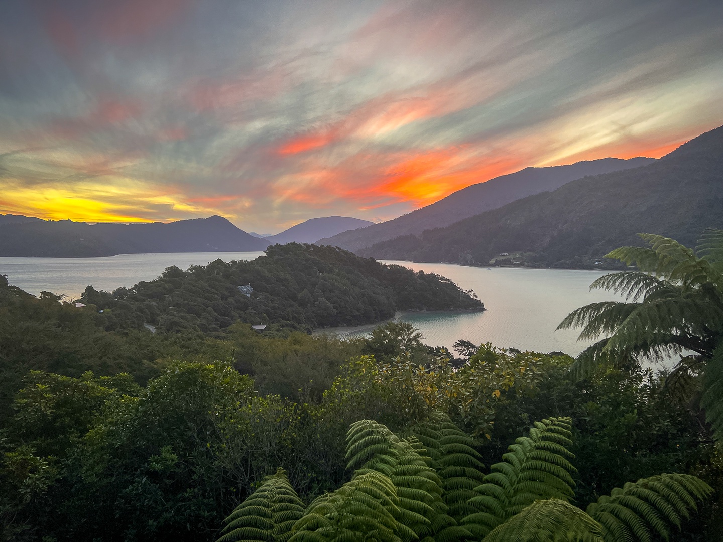 Dramatic sunset paints the sky in vibrant oranges and yellows over pristine lake waters surrounded by lush mountain ranges and tropical vegetation.