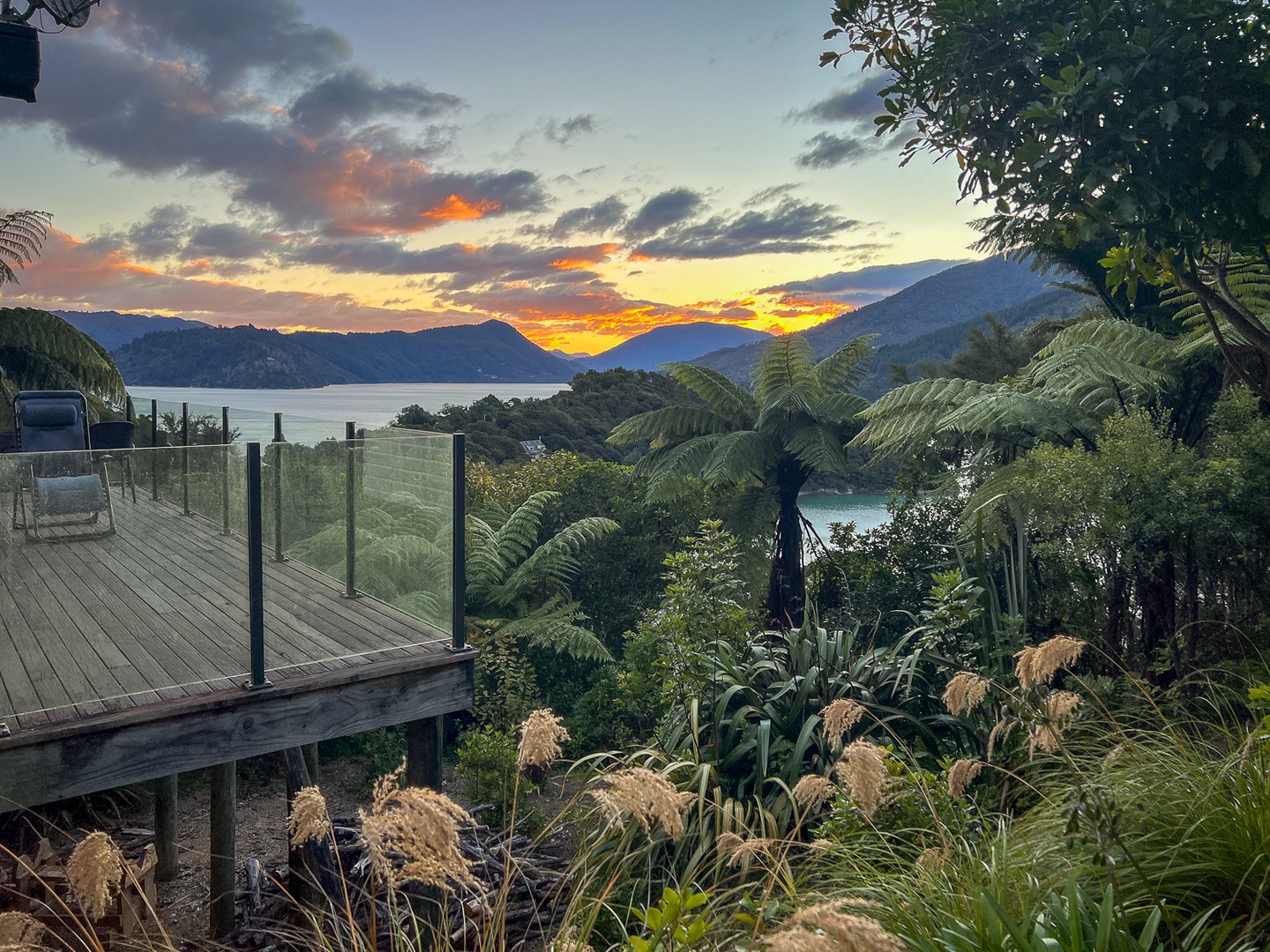 Stunning lakeside deck overlooking dramatic mountain ranges and native forest, with vibrant sunset colors painting the evening sky.